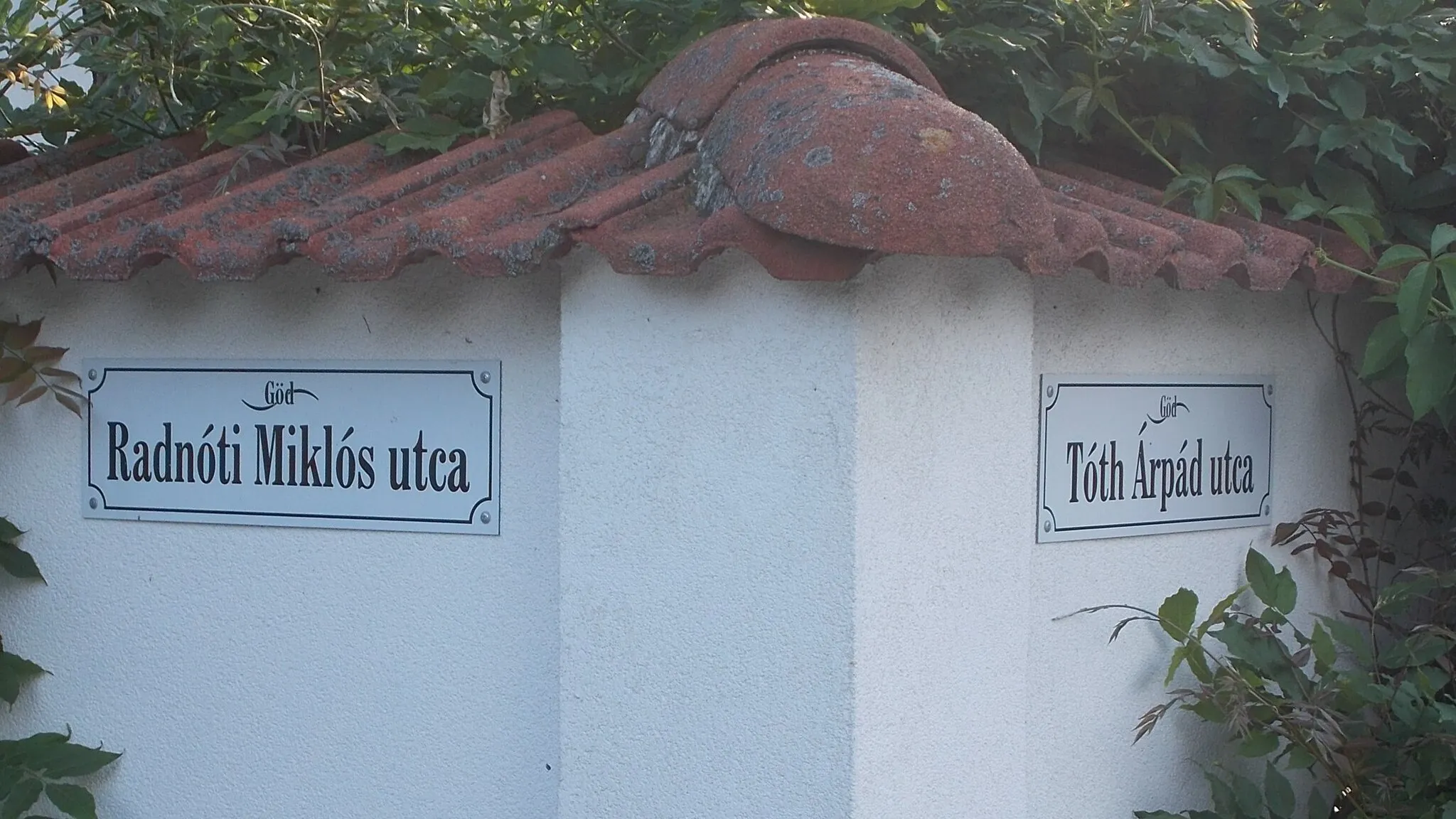 Photo showing: : Street name sign, tile covered fence wall - Radnóti Miklós Street and Tóth Árpád Street, Alsógöd neighborhood, Göd, Pest County, Hungary.