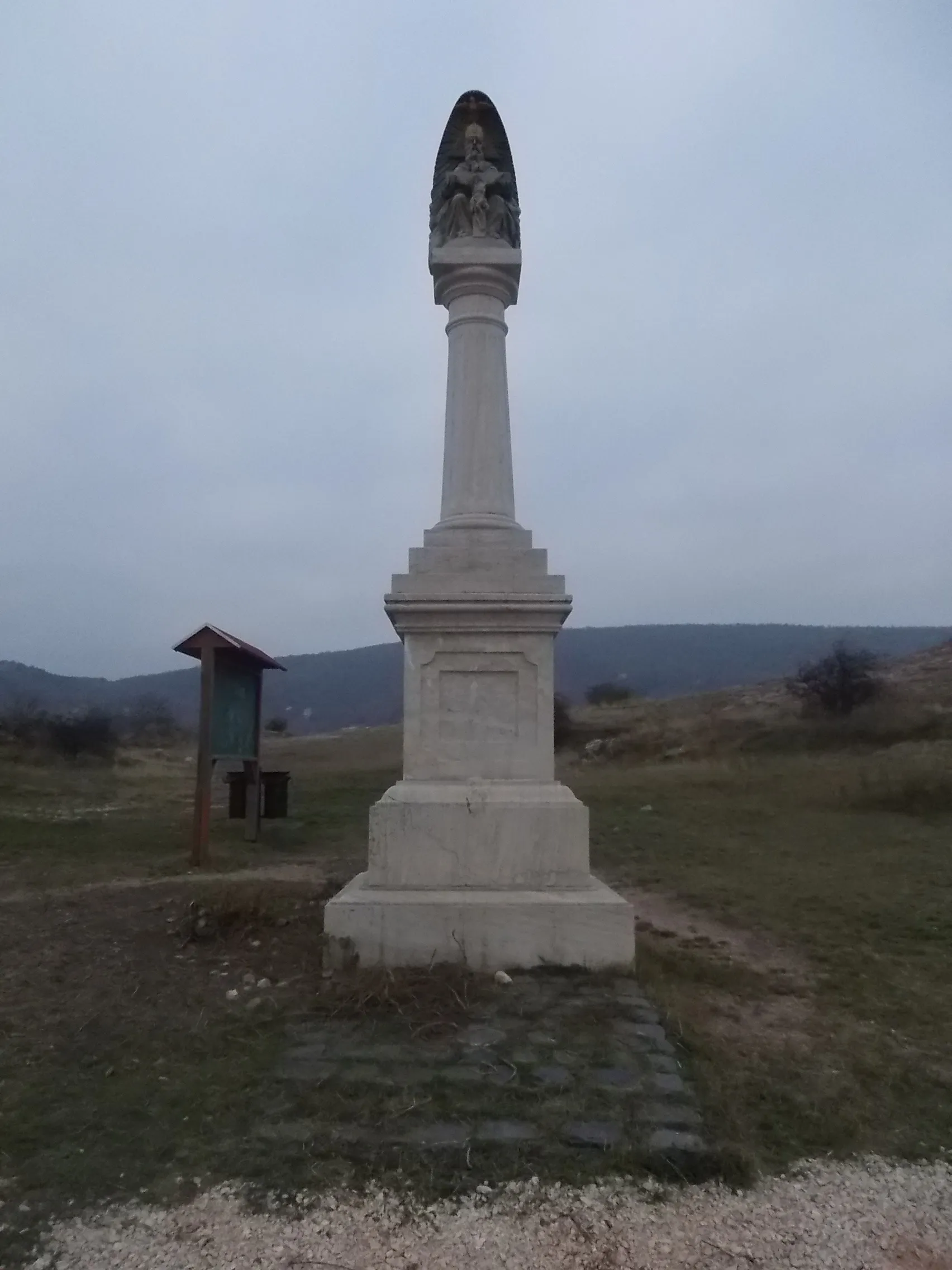 Photo showing: Holy Trinity column by Péter Rákos (installed in 2004). Sitting God the Father staue hold a crucifix at back dove/holy trinity relief. Next to the hill chapel on the ~Stone Hill. It is part of the Stone Hill Religious Complex: stone crucifix, chapel and Holy Trinity column, and part of the ~Stone Hill Nature Protected Area (under local grade level protection) - Historical city center neighborhood, Budaörs, Pest County, Hungary.