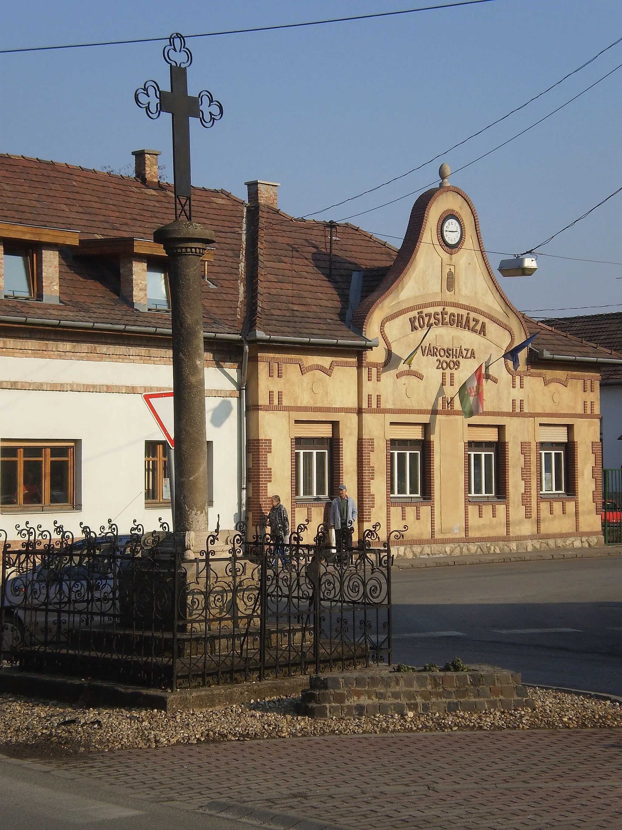 Photo showing: Budakalász, Petőfi tér: városháza és műemlék kereszt ID 6918, Vaskereszt, klasszicista, 1841. Vasráccsal körülvett négy kőlépcsőn áll, négyzetes talapzata ezen elöl 1841-es évszám. Ezen emelkedik a négy gömbön nyugvó, csigákkal díszített lábazaton a simatörzsű oszlop, melynek tányérja alatt fogrovatos öv fut körbe.  Az oszlop tetején karéjos szárain áttört kovácsoltvas kereszt áll..