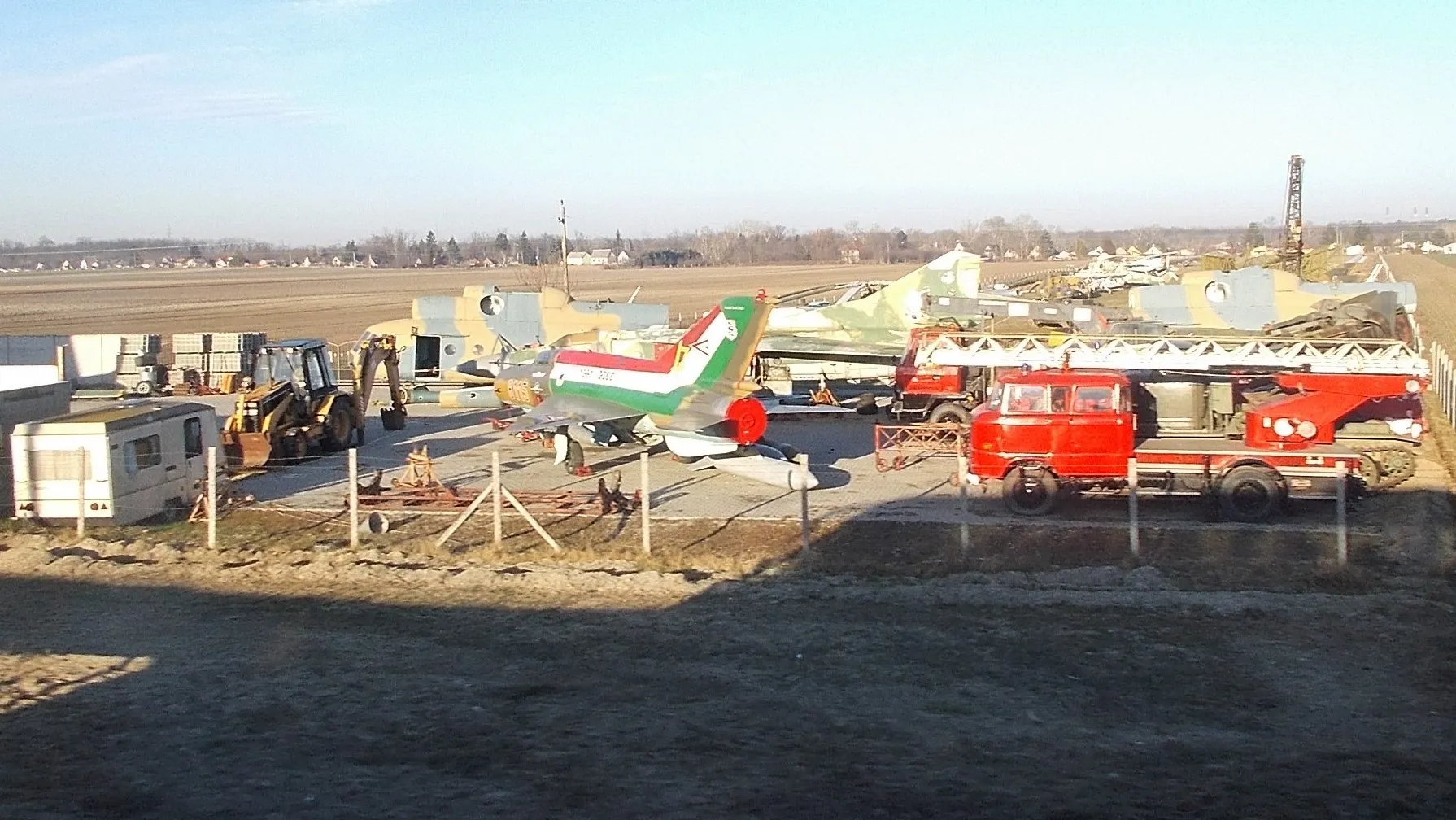 Photo showing: : Vintage (scrapped) aircrafts and other vehicles. In front MiG and Mi-8A and IFA fire truck. Photo taken through the bus window. - Dunavarsány, Pest County, Hungary.