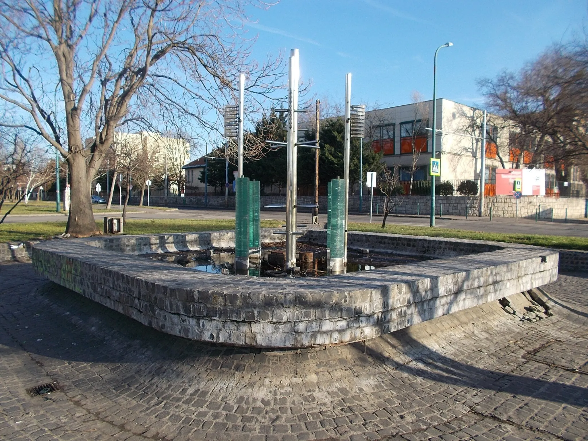 Photo showing: : Nonfigurative fountain by Zoltán Bohus, Béla Hámori (1979 works, the pool of the fountan built cobblestone, middle of the pool is an abstract green glass and steel, five meters high, illuminated). A long cylinder stand in the middle it is creates a regular glass-like shape from the water,  four concave plates and four tubes create four convex watersurfaces...Saddly it did not work. On left is a part of the Hungarian Dance Academy (HDA) Campus - Located in a small Park at Mexikói Street, Herminamező neighbourhood, Budapest District XIV.