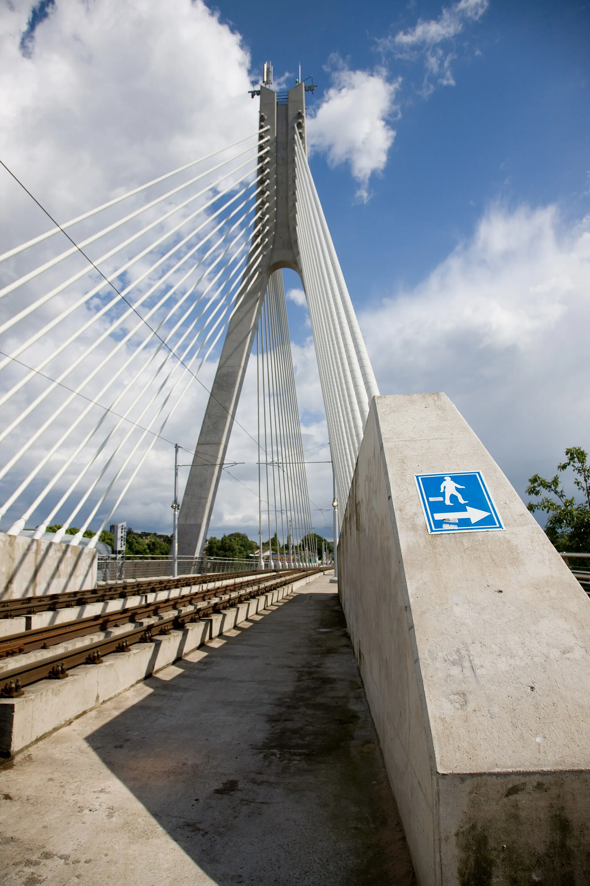 Photo showing: The 2000s saw two major developments relating to transport which affected Dundrum greatly. Firstly, a road bypass was built behind the west side of the village, taking most of the traffic away from the main street. Secondly, the old railway line has been converted to a tram line, running the new Luas tram system. Since the railway embankment was cleared away in the 1970s to make room for a major road junction at the north end of the village, the tramway designers now had the problem of getting the trams across a very wide crossing without any place for supporting pillars.

The solution was innovative: a suspension bridge.