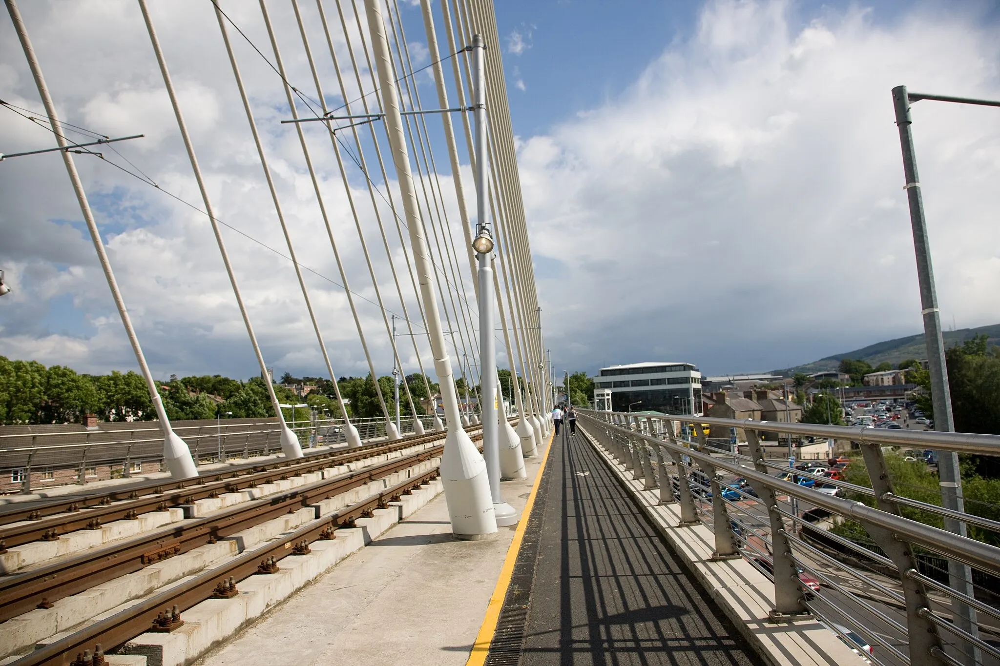 Photo showing: The 2000s saw two major developments relating to transport which affected Dundrum greatly. Firstly, a road bypass was built behind the west side of the village, taking most of the traffic away from the main street. Secondly, the old railway line has been converted to a tram line, running the new Luas tram system. Since the railway embankment was cleared away in the 1970s to make room for a major road junction at the north end of the village, the tramway designers now had the problem of getting the trams across a very wide crossing without any place for supporting pillars.

The solution was innovative: a suspension bridge.