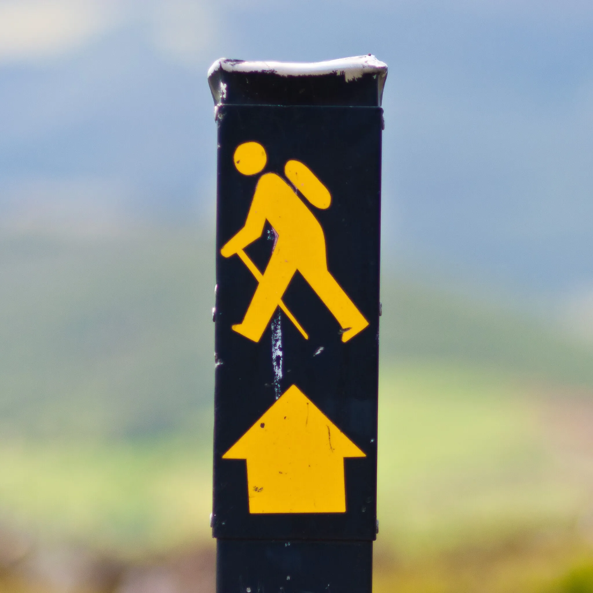 Photo showing: Waymarking sign, comprising an image of a walking man and a directional arrow in yellow, used in Ireland to denote a National Waymarked Trail. The design was copied from the symbol used to waymark the Ulster Way in Northern Ireland and has since become the standard waymarking image used for long-distance trails in the Republic of Ireland.