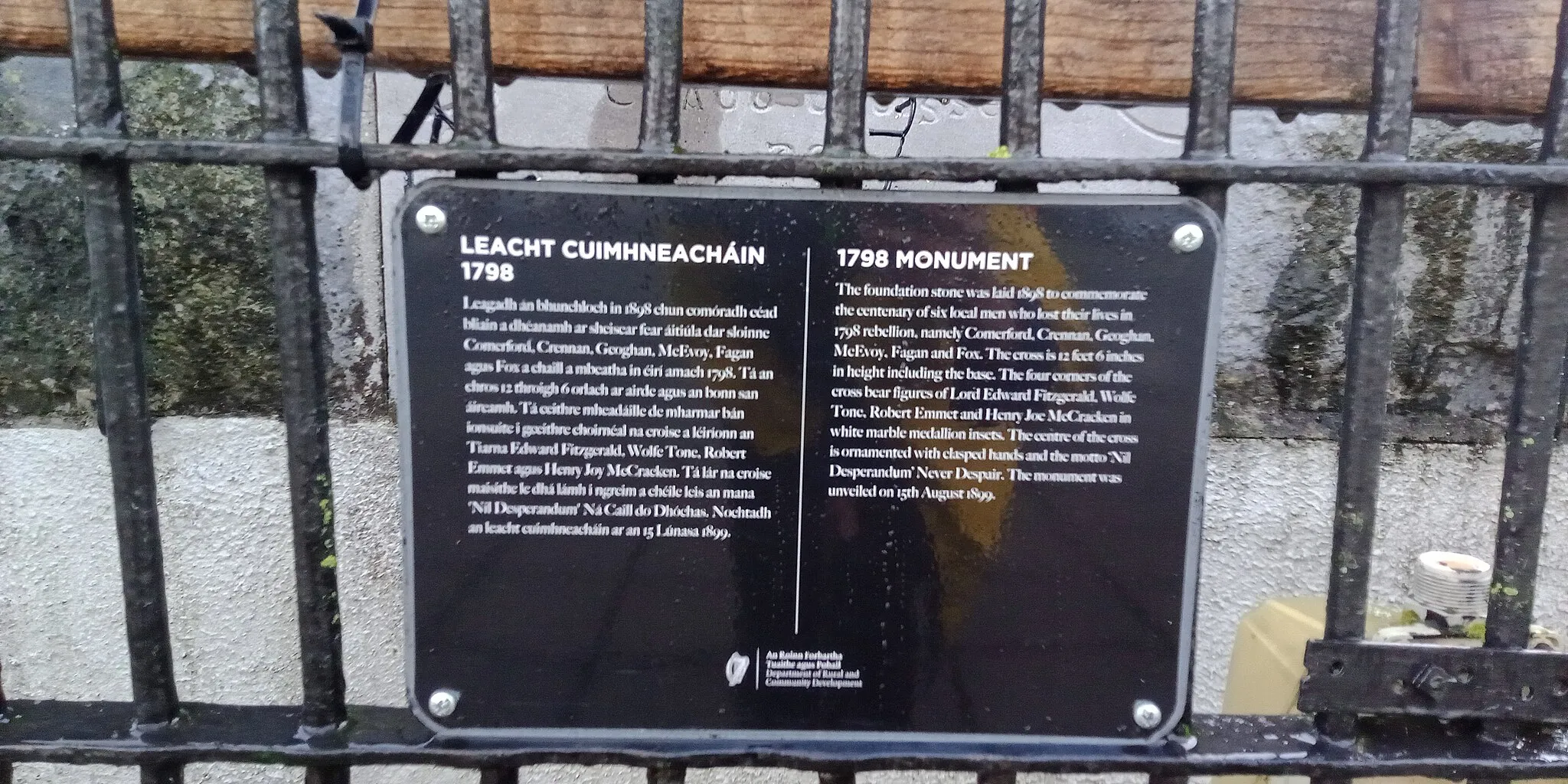 Photo showing: bilingual sign on the 1798 Monument in Ballinakill. The English text reads "1798 monument. The foundation stone was laid 1898 to commemorate the centenary of six local men who lost their lives in 1798 rebellion, namely Comerford, Grennan, Geoghan, McEvoy, Fagan and Fox. The cross is 12 feet 6 inches in height including the base. The four corners of the cross bear figures of Lord Edward Fitzgerald, Wolfe Tone, Robert Emmet and Henry Joe McCracken in white marble medallion inlets. The centre of the cross is ornamented with clasped hands and the motto "Nil Desperandum" Never Despair. The monument was unveiled on 15th August 1899."