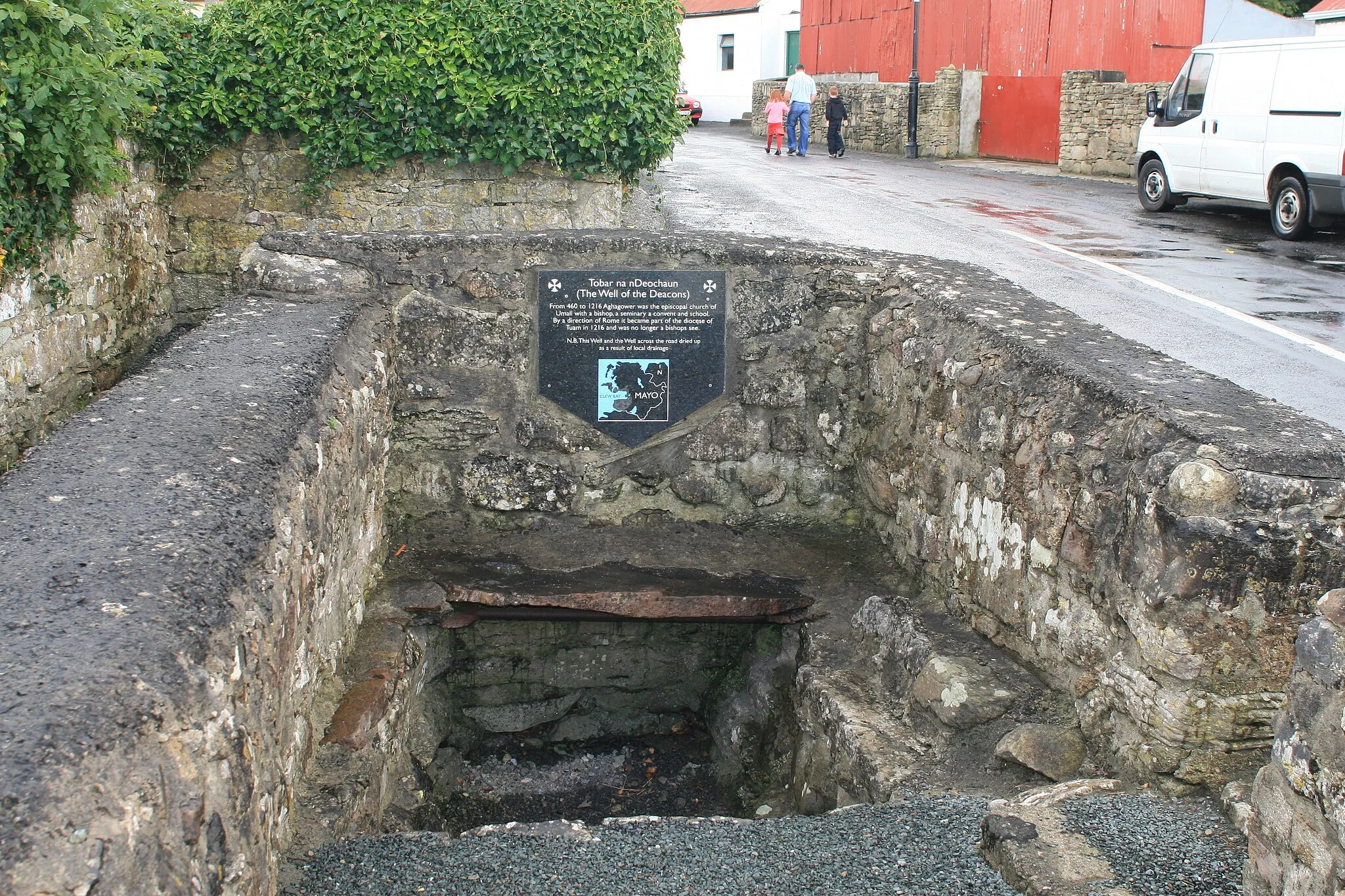 Photo showing: One of the two holy wells at Aghagower. The depicted plate reads: Tobar na nDeochaun (The Well of the Deacons): From 460 to 1216 Aghagower was the episcopal church of Umall with a bishop, a seminary a convent and school. By a direction of Rome it became part of the diocese of Tuam in 1216 and was no longer a bishops see. N.B. This Well and the Well across the road dried up as a result of local drainage.