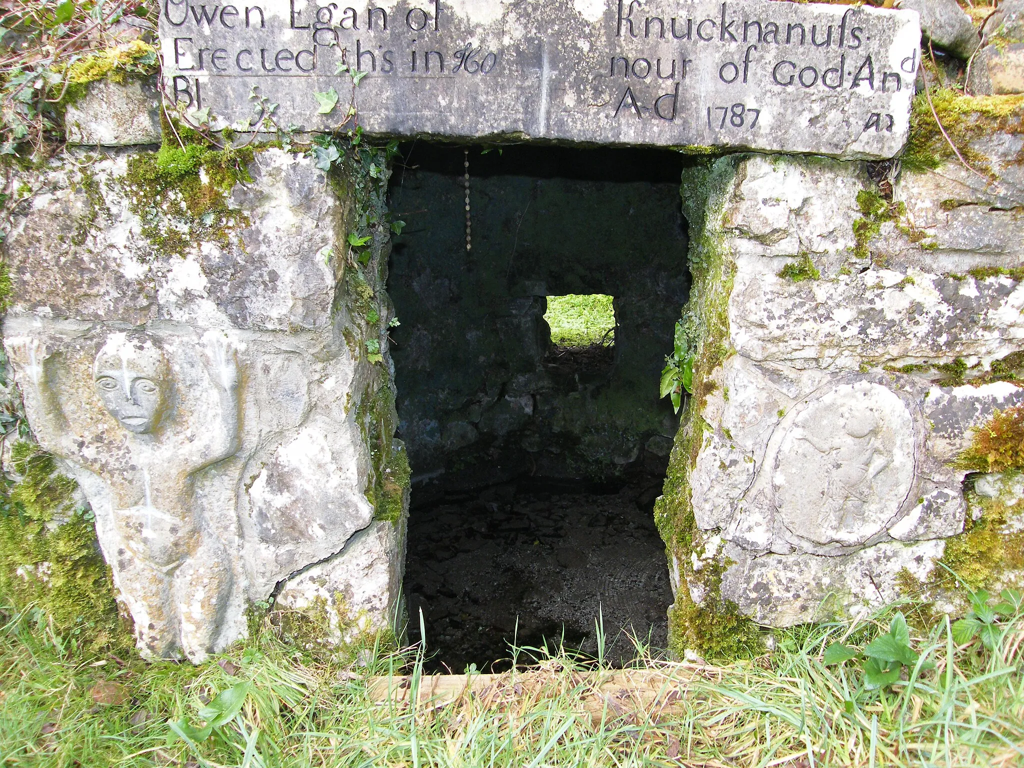 Photo showing: Entrance to St. Bridget's Well at Castlemanger, Co. Cork, Ireland.  A figure somewhat similar to a sheela-na-gig except for hand position and legs not spread is to left of entrance; a smaller figure is to the right; and a 1787 inscription is above.