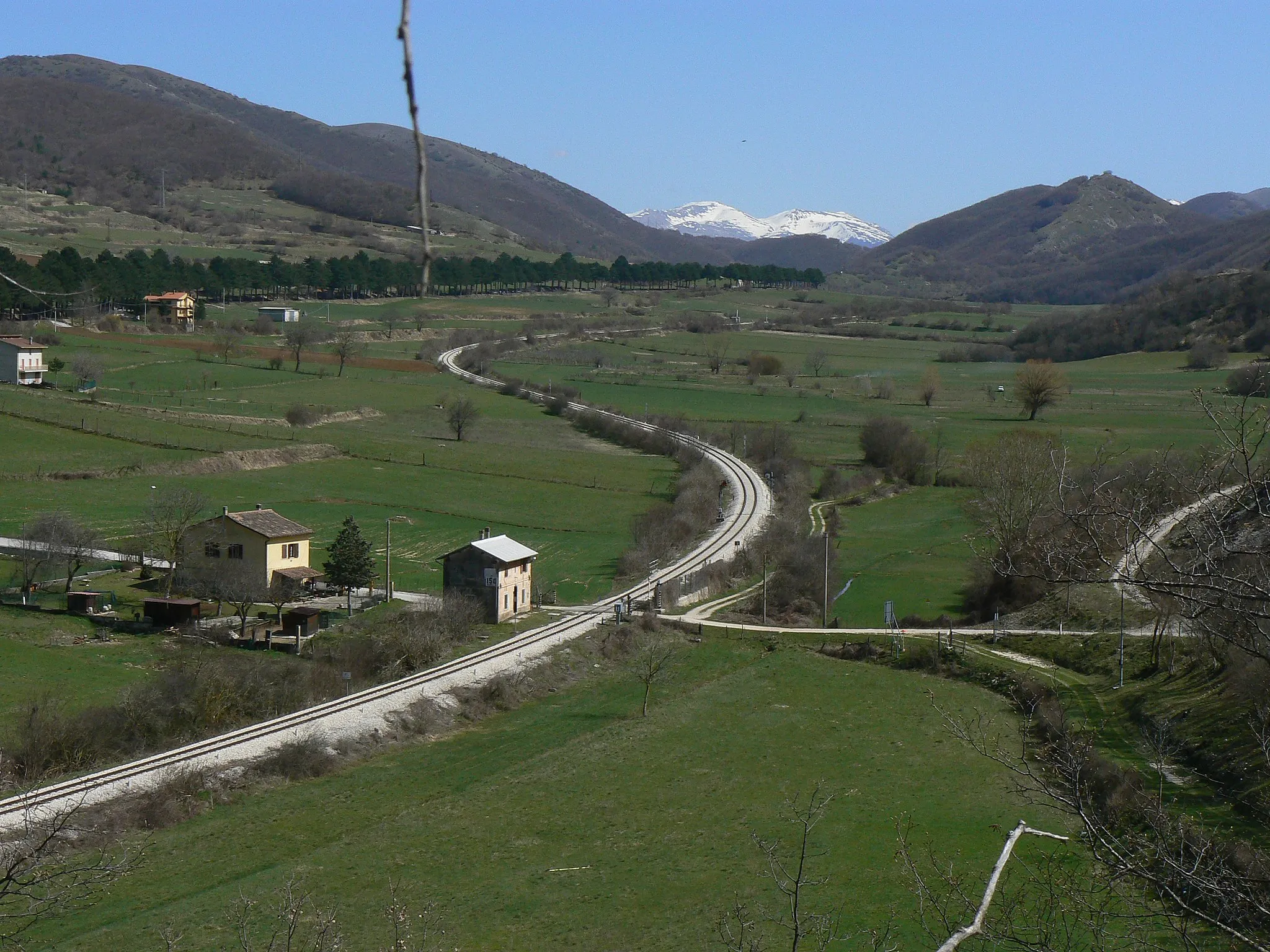 Photo showing: View of Sella di Corno plateau, in the point where lies the border between Lazio and Abruzzo, Italy. The tree-lined street at left is state highway n. 17, while on foreground Terni-Rieti-L'Aquila railway can be seen (trait between Rocca di Corno station and Sella di Corno station, which is the highest point of the line at 989 metres on the sea level) with a level crossing without barriers and a watchman's house indicating the kilometer 150.