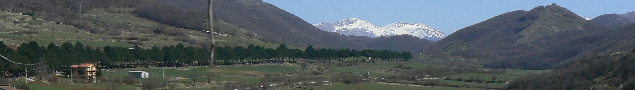 Photo showing: View of Sella di Corno plateau, in the point where lies the border between Lazio and Abruzzo, Italy. The tree-lined street at left is state highway n. 17, while on foreground Terni-Rieti-L'Aquila railway can be seen (trait between Rocca di Corno station and Sella di Corno station, which is the highest point of the line at 989 metres on the sea level) with a level crossing without barriers and a watchman's house indicating the kilometer 150.