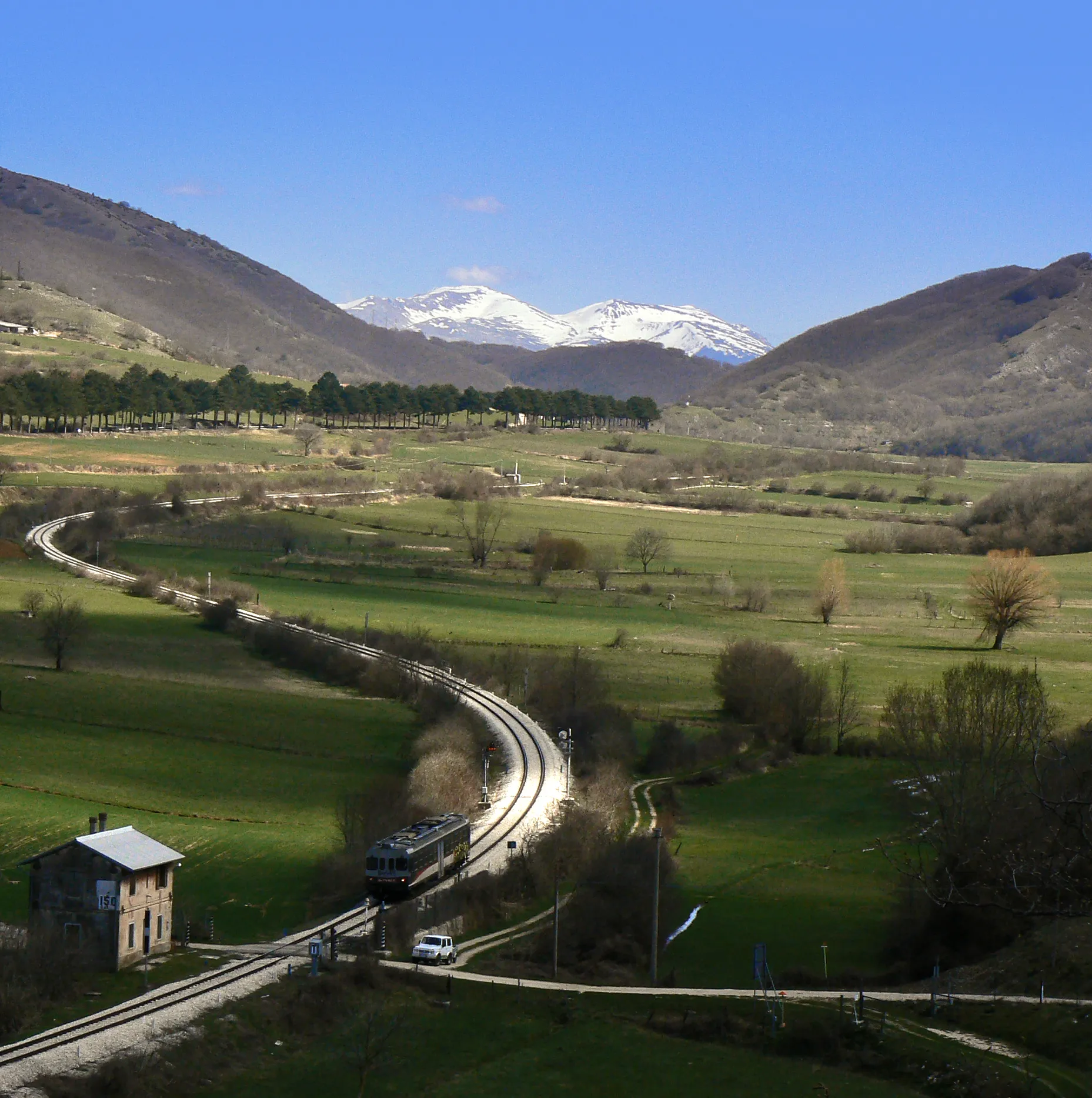 Photo showing: View of Sella di Corno plateau, in the point where lies the border between Lazio and Abruzzo, Italy. The tree-lined street at left is state highway n. 17, while on foreground Terni-Rieti-L'Aquila railway can be seen (trait between Rocca di Corno station and Sella di Corno station, which is the highest point of the line at 989 metres on the sea level) with a level crossing without barriers and a watchman's house indicating the kilometer 150, with an ALn 776 class diesel multiple unit owned by Ferrovia Centrale Umbra passing through.