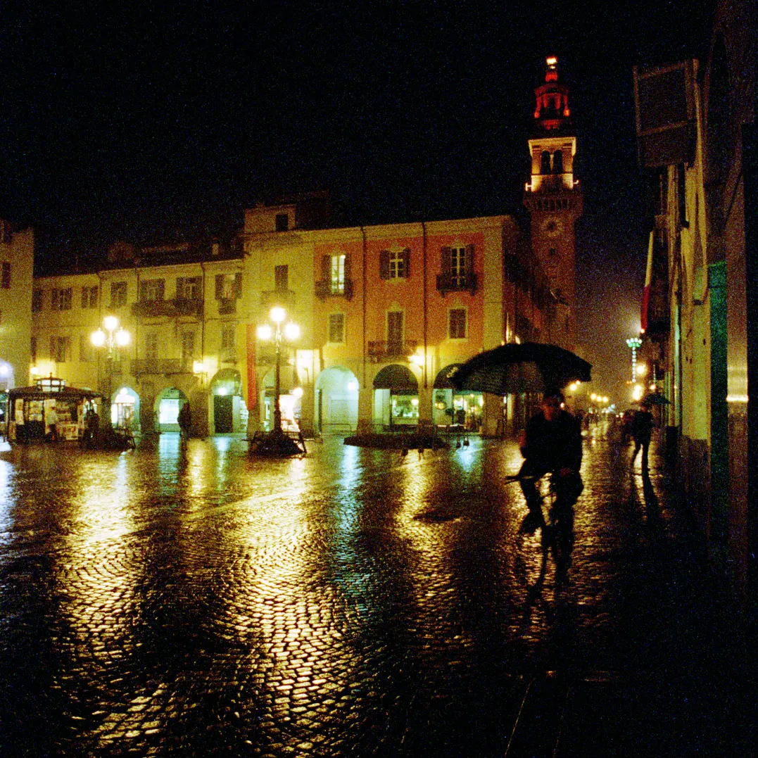 Photo showing: Casale Monferrato: Piazza Mazzini after dark. In the backgound Via Saffi leads past the Torre Civica, while the arcades on the left mark the beginning of Via Roma. Cyclist with umbrella in foreground.