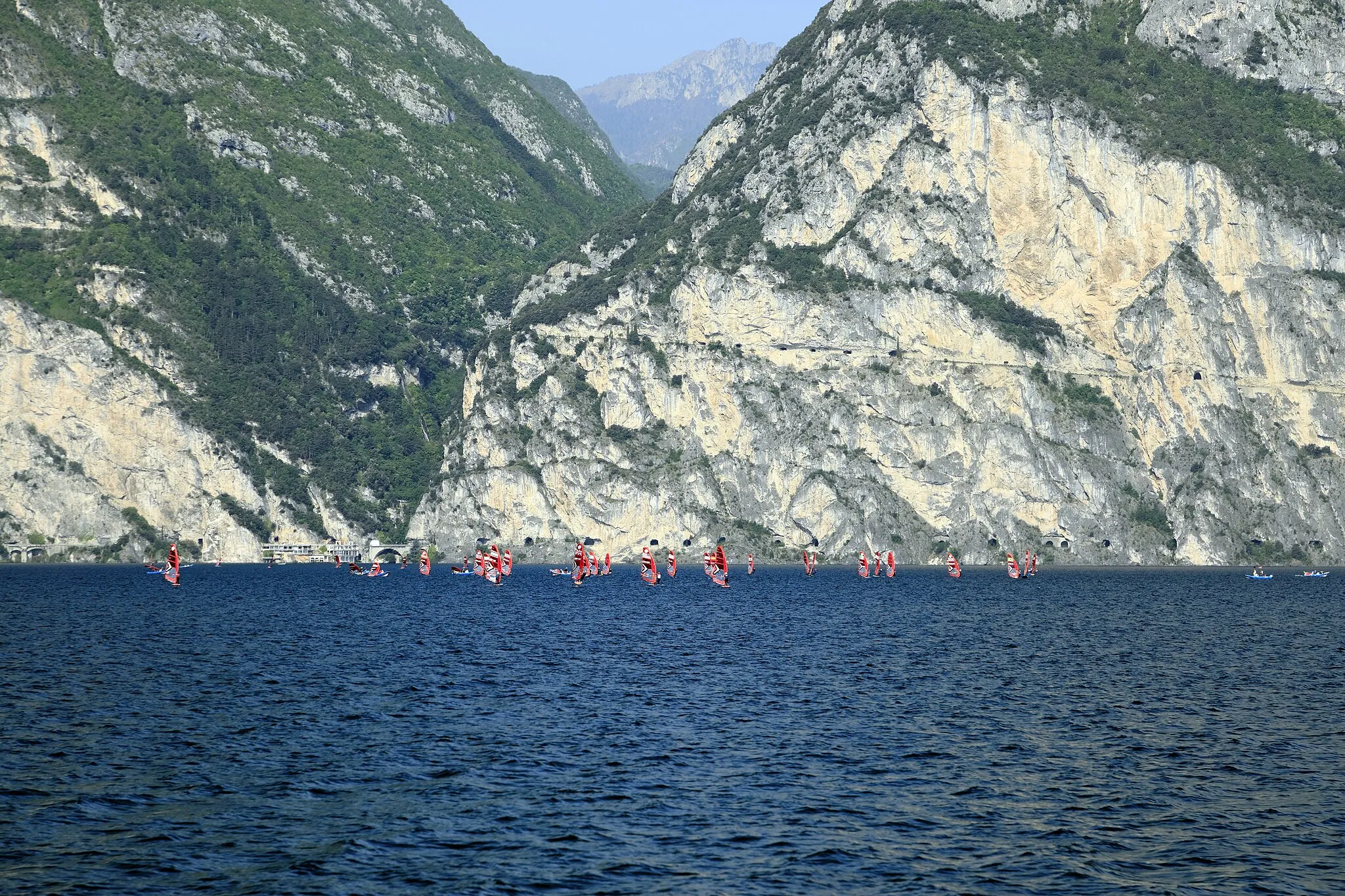 Photo showing: Vom Kursschiff Trento am Anleger Torbole Richtung Westen; durch den berechenbar gleichmäßigen Wind eignet sich der Gardasee für die Seglerausbildung. Zum Aufnahmezeitpunkt nutzt es eine regelrechte Surferflotille mit Ostnordostkurs.
Auf gleichmäßiger Höhe verläuft am Westufer die tunnelreiche Uferstraße SS 45bis Gardasena Occidentale zwischen Riva und Gargnano. Darüber ansteigend in die Felswand eingebaut führt die alte Ponalestraße ins Ledrotal. Sie war bis 1998 die einzige Verbindung ins Ledrotal, wurde dann durch die neue SS 240 mit einem langen Tunnel ersetzt und für jeden Verkehr gesperrt. Seit einer Instandsetzung 2004 ist sie jedoch für Fahrradfahrer und Fußgänger wieder nutzbar.