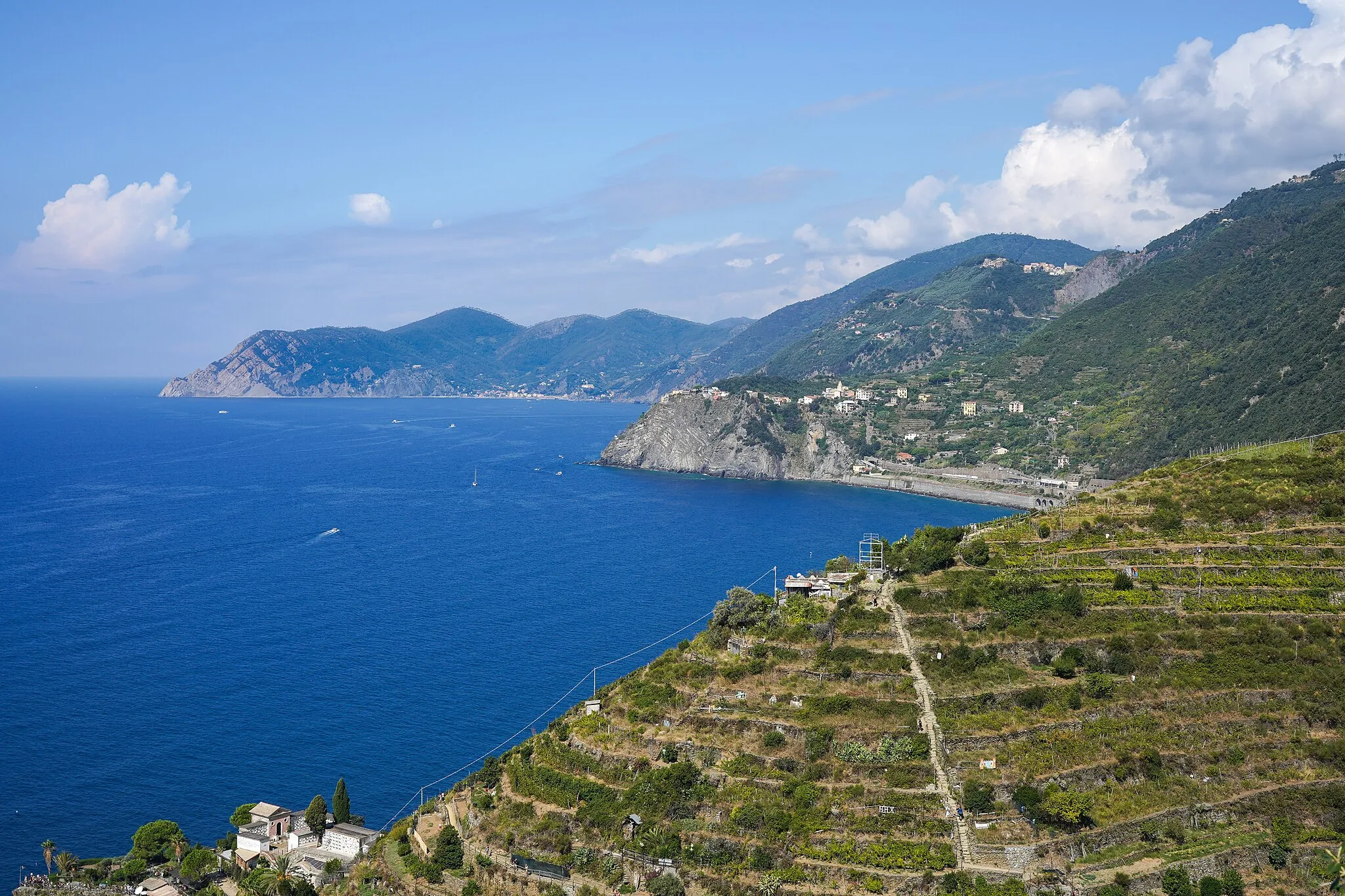Photo showing: View of coastline NW from above Manarola: Manarola cemetery and fields and steep trail to Volastra in foreground, Corniglia Station and town centre, San Bernardino above, Monterosso al Mare and Punta Mesco.  Depicts the north half of Cinque Terre National Park coastline, about 10 km (6.2 mi) out of 20 km (12 mi). Cinque Terre, Liguria, Italy