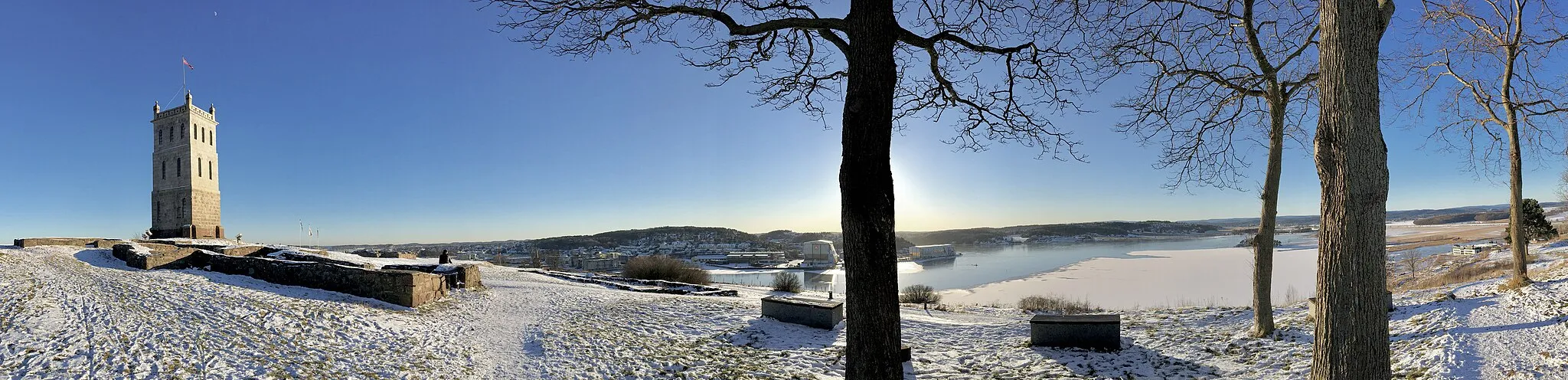 Photo showing: The Slottsfjellet, a hill located in central Tønsberg,Norway, with the Slottsfjelltårnet tower from 1888 and the ruins of the medieval Tunsberghus Fortress (Tunsberghus festning). View towards the small city beneath the small mountain, the Byfjorden/Tønsbergfjorden fjord, Kaldnes, Hogsnes, Stokkelandet, Ilene, Jarlsberg, Sem, Frodeåsen, Solvang, the Tønsberg Stasjon railway station from 1915, Peder Lagmanns gate, the Vestfold sentralsykehus hospital, Kilen, and other parts of the surrounding landscape in Vestfold by the Oslofjorden fjord. An old farm house (Vestfoldtunet) and a stabbur on Tallak, as part of the Slottfjellsmuseet museum. Distorted panorama photograph taken on February 15th, 2016, with the low hanging afternoon sun on a clear, blue sky making long shadows on the snowy ground.