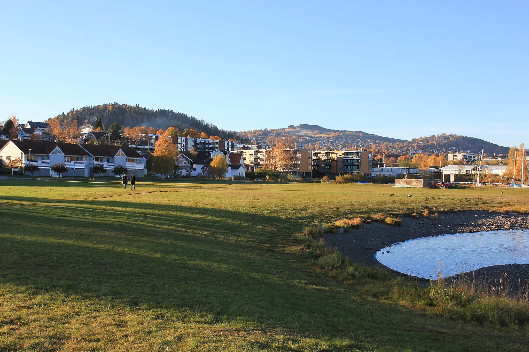 Photo showing: Gjøvik er toppenes by. Til venstre ser vi Hovdetoppen sentralt i byen, røyken i forgrunnen er vanndamp fra Hunton. Midt i bildet ligger Bergstoppen, hvor det hvert år arrangeres et toppløp opp fra sentrum. Til høyre er Tranbergtoppen, denne kommer best til sin rett når man ankommer byen nordfra. I forgrunnen ses gressletta i Fredevika.