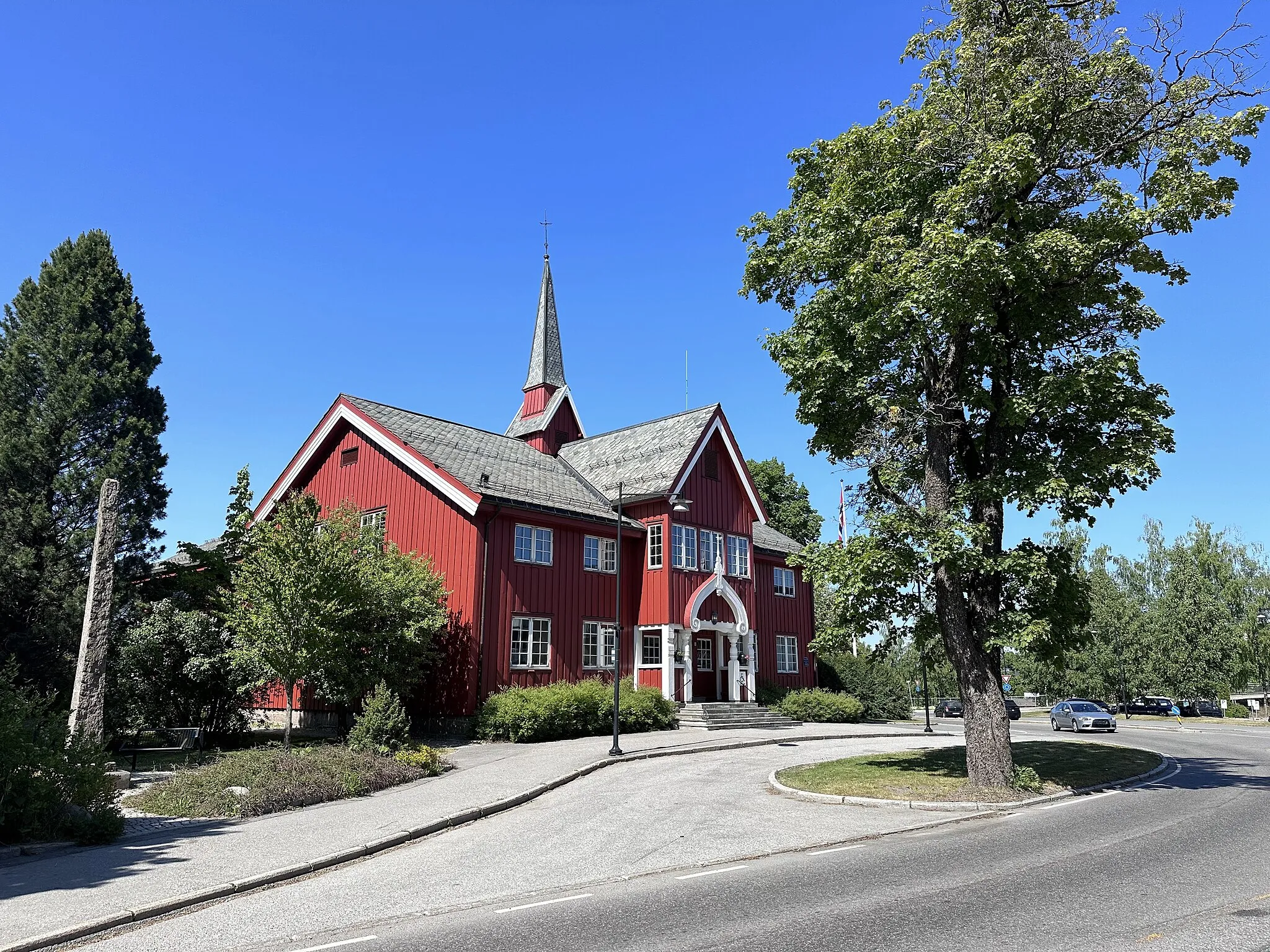 Photo showing: Herredshuset ('the town hall' or 'the municipal building') in Jessheim, Ullensaker municipality, Norway. Herredshuset is a red-painted wooden building designed by the architect Holger Sinding-Larsen, and opened 1901 as a municipal building, featuring a banquet hall and spaces for the local bank. The building functioned as the municipal offices until 1967. It now serves as a venue for courses and events. Near the Herredshuset is a five-meter-high monument stone erected on May 17, 1914, "in memory of the soldiers from Ullensaker in the War of 1814 – Lier and Matran" and a World War II memorial plaque "in memory of those from Ullensaker who fell in the fight for Norway's freedom 1940-1945."