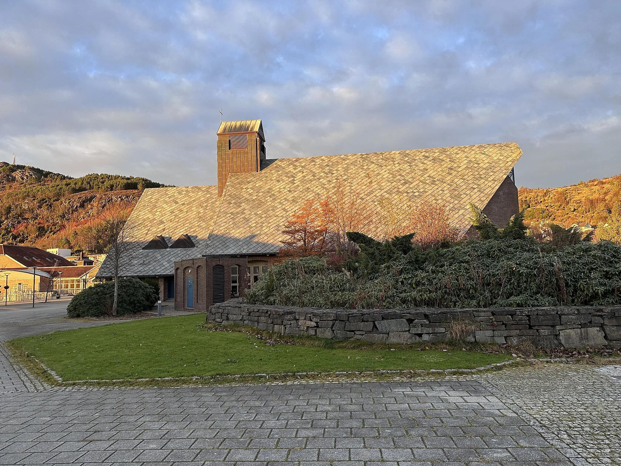 Photo showing: Aksdal Church in Tysvær municipality in Rogaland county, Norway. The church was designed by architect Stein Jarle Helgeland and was consecrated in 1995. The brick building is located in the center of Aksdal, with Aksdal shopping center and Tysværtunet cultural center, care home, swimming facility, and other amenities right nearby. The picture was taken on a November afternoon with warm light and long shadows from the low sun that is about to set.