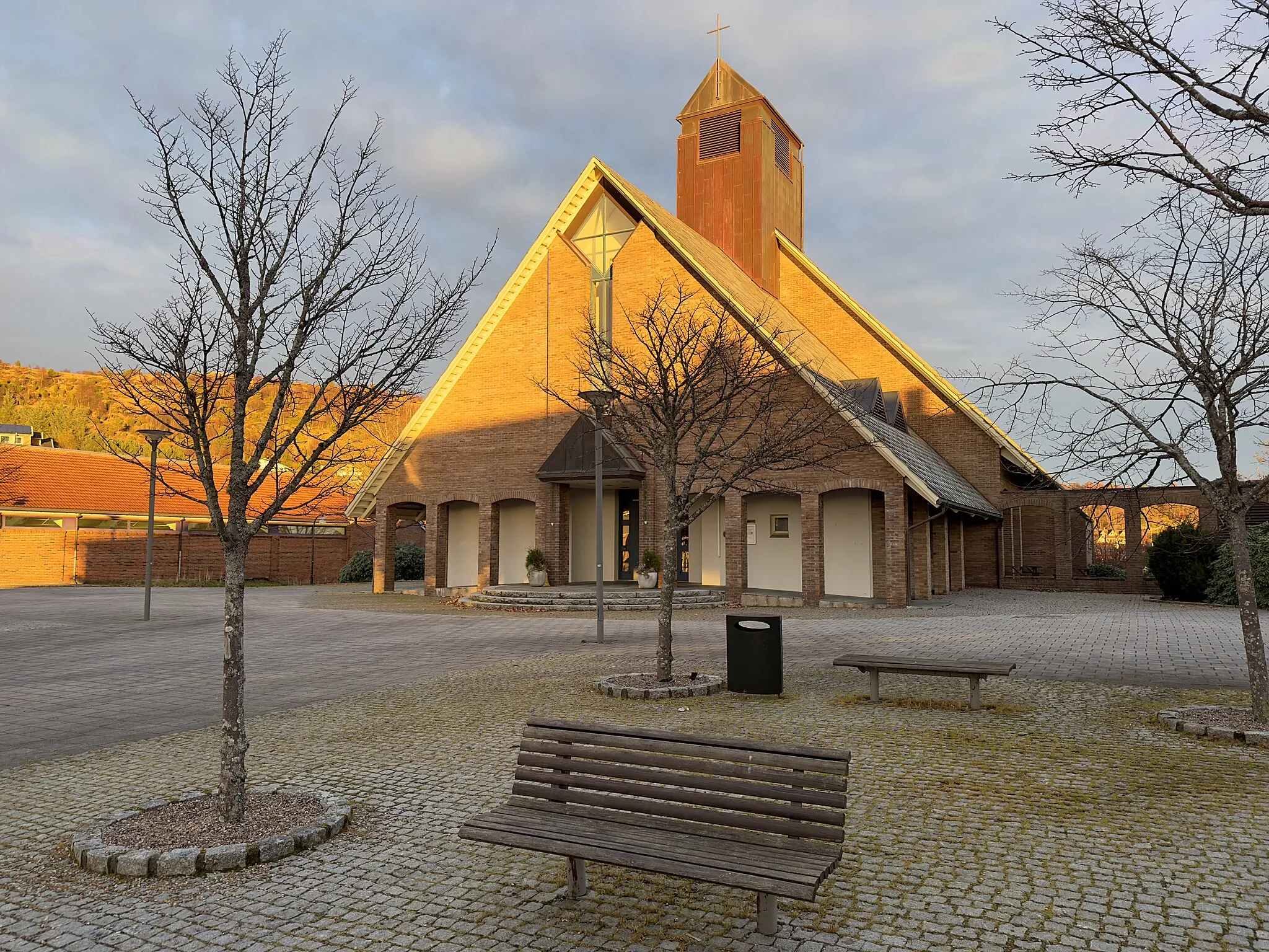 Photo showing: Aksdal Church in Tysvær municipality in Rogaland county, Norway. The church was designed by architect Stein Jarle Helgeland and was consecrated in 1995. The brick building is located in the center of Aksdal, with Aksdal shopping center and Tysværtunet cultural center, care home, swimming facility, and other amenities right nearby. The picture was taken on a November afternoon with warm light and long shadows from the low sun that is about to set.