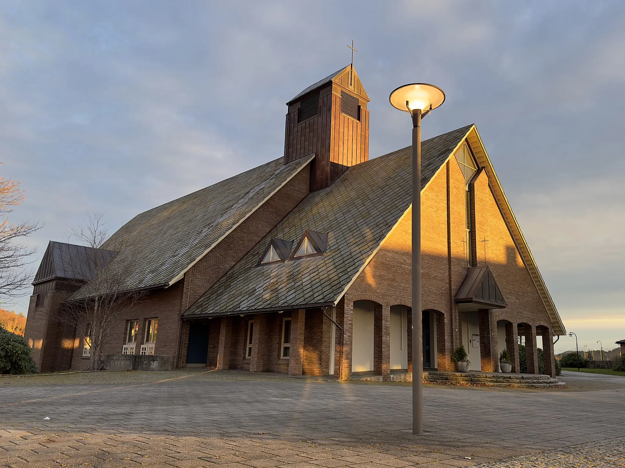 Photo showing: Aksdal Church in Tysvær municipality in Rogaland county, Norway. The church was designed by architect Stein Jarle Helgeland and was consecrated in 1995. The brick building is located in the center of Aksdal, with Aksdal shopping center and Tysværtunet cultural center, care home, swimming facility, and other amenities right nearby. The picture was taken on a November afternoon with warm light and long shadows from the low sun that is about to set.