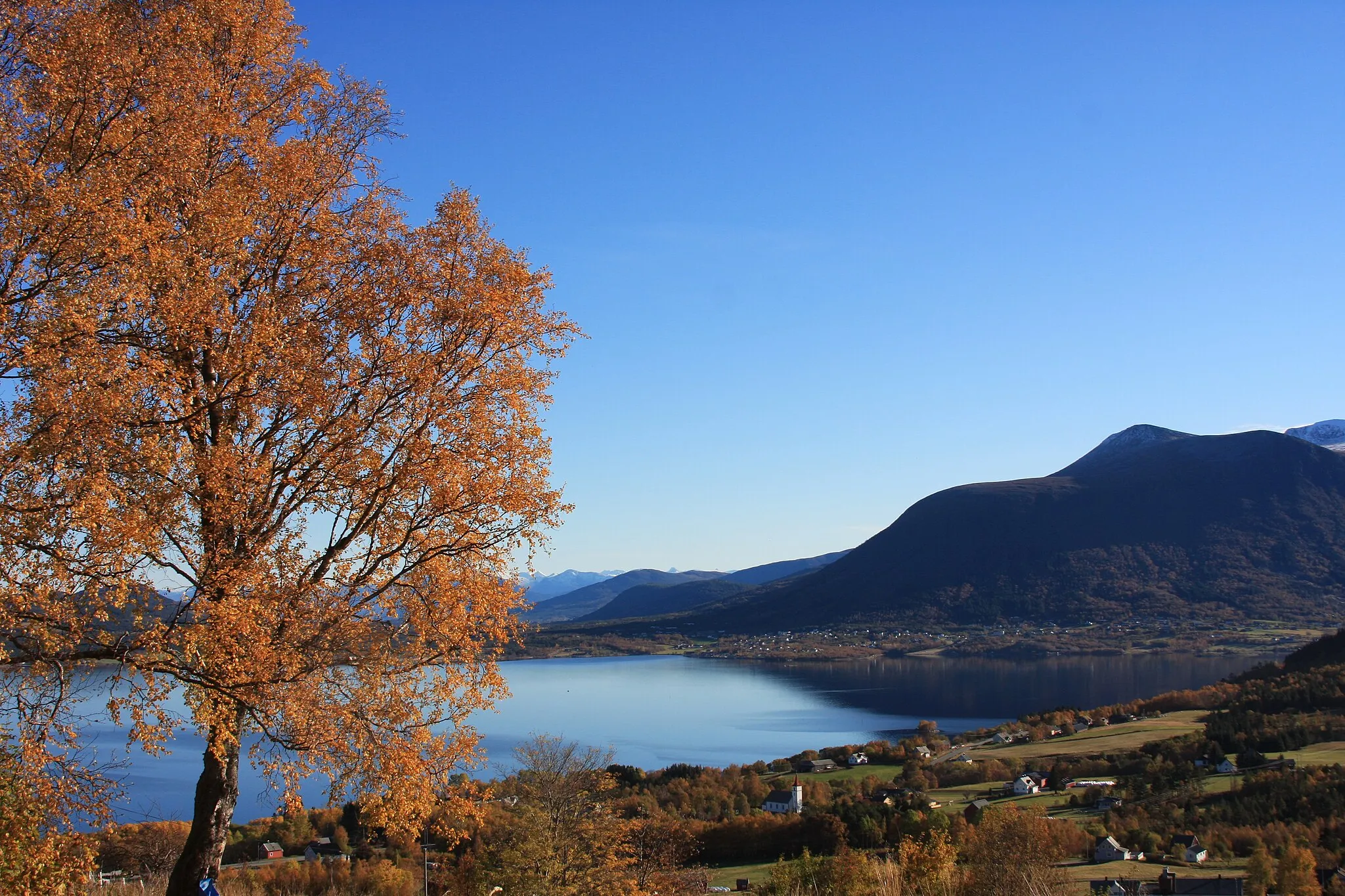 Photo showing: Panorammic view in autumn colors over Fiksdal, a village in the county Møre og Romsdal in Norway. You see the white church in the middle of the village and the fjord Tomrefjord.