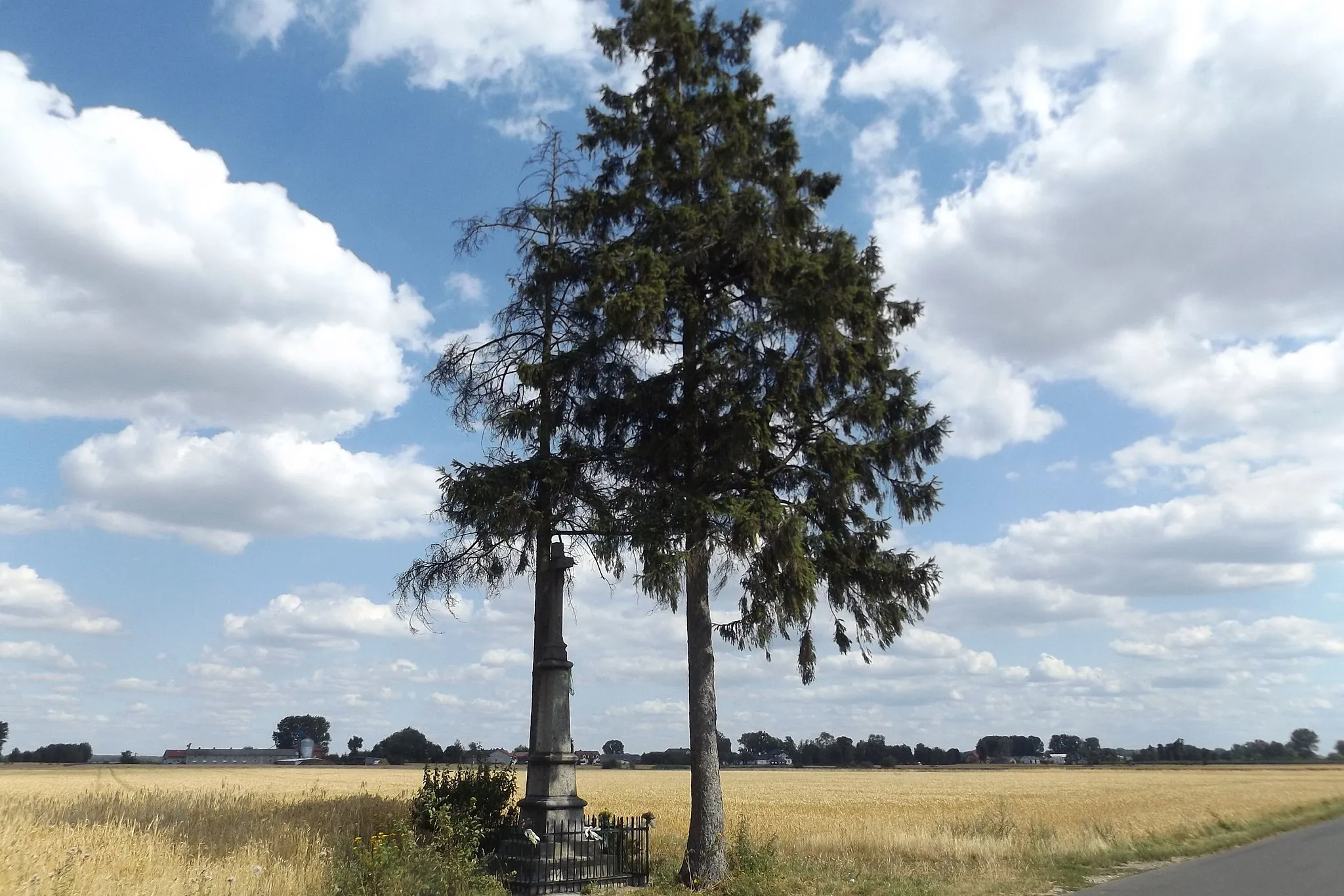 Photo showing: Wayside shrine located in Osiek between parts of Osiek Drugi and Osiek Trzeci; on the NE side of the intersection of the road to Łowicz with the green lane leading from Wejsce to Osiek Drugiego; in 2019. In the background Osiek Trzeci (east part of village Osiek).