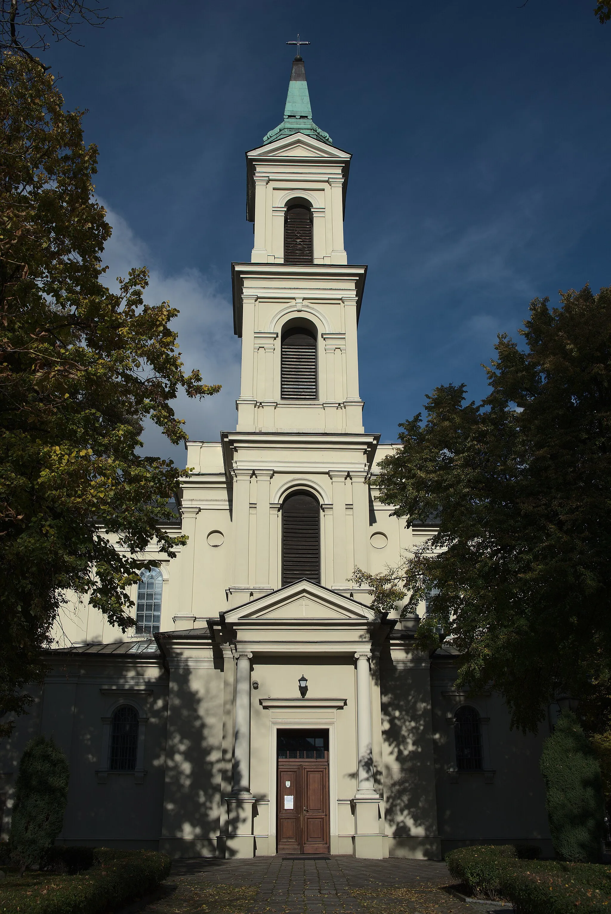 Photo showing: Front kościoła świętego Wojciecha w Kielcach. Budynek jest biały z lewej oraz prawej widać dwa drzewa które rzucają cień na budynek. Sam budynek pojedynczą wieżę, która ma trzy piętra, każde piętro ma pojedyncze okno przez które nic nie widać. Na parterze kościoła znajdują się wejściowe drzwi. Parter oraz pierwsze piętro są szersze mają jeszcze zabudowania po prawej i po lewej. Natomiast drugie i trzecie piętro to już sama wieża nie wiele szersza niż samo okno.