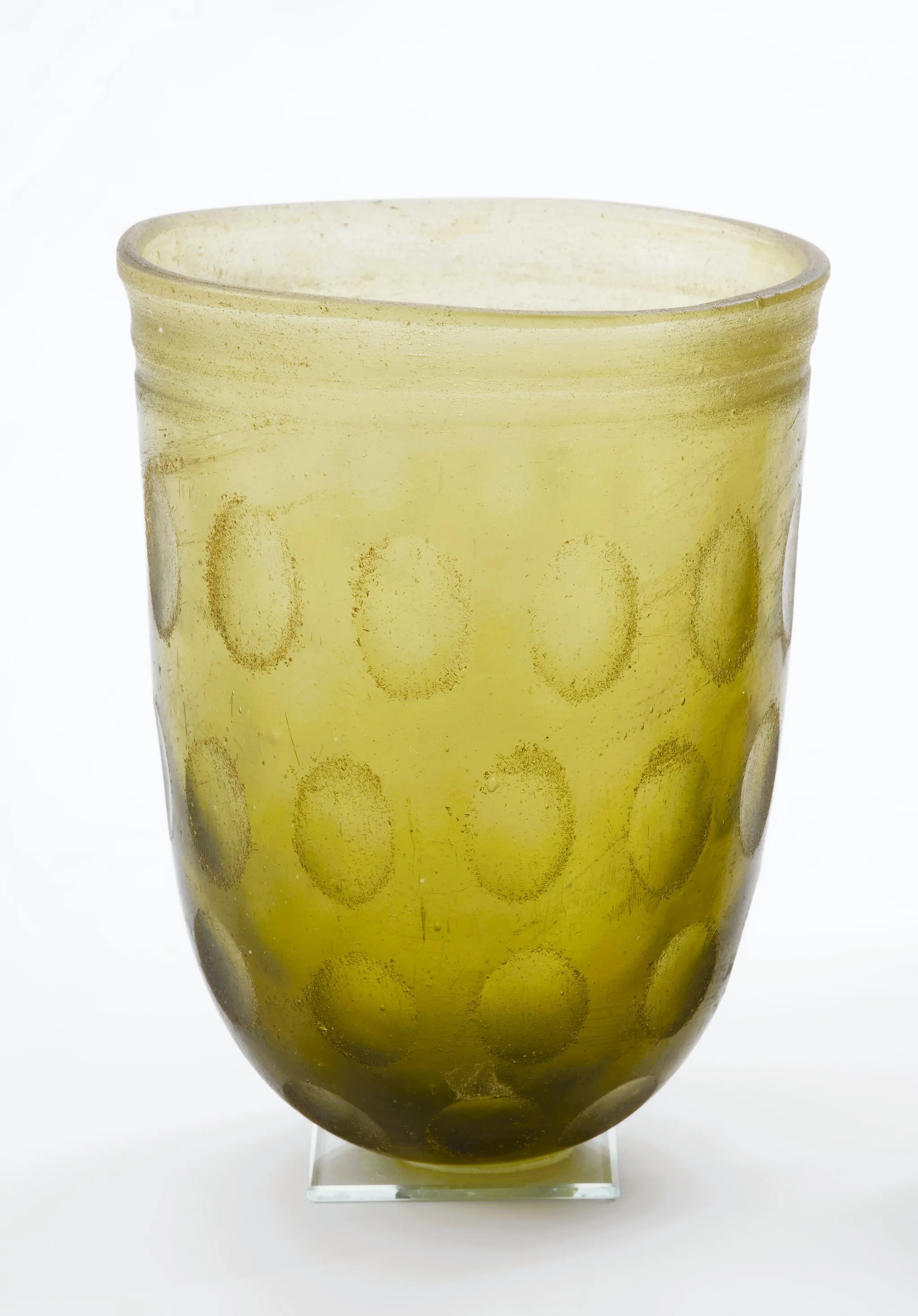 Photo showing: MNS/A/22211 The glass vessel (beaker) with thick walls, is light green and decorated with polished ovals, arranged in a system of four stripes running around the vessel, and two lines engraved under the edge of the rim. The glass is transparent. It belongs to a group of glass vessels with a characteristic form and equally typical ornamentation, known in the literature as “Kowalki type” vessels. The name comes from another location in Western Pomerania.