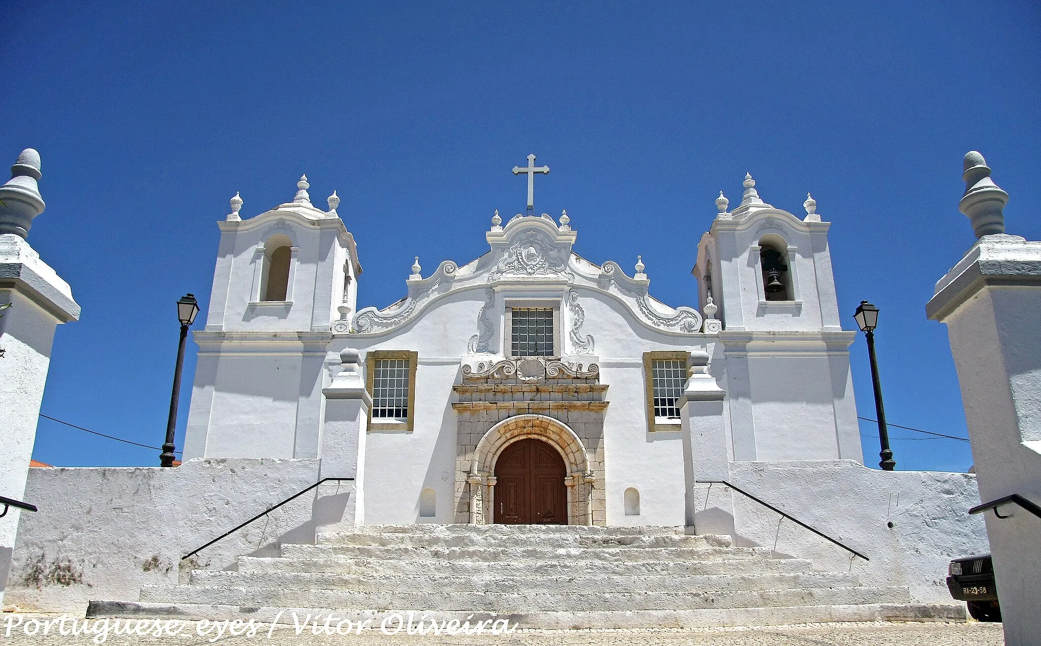 Photo showing: De remota fundação, a Igreja Matriz da bonita povoação de Estômbar, que integra o concelho da Lagoa, foi remodelada no reinado de D. Manuel I. No entanto, o século XVIII havia de a reformar quase totalmente, conservando este templo ainda alguns sinais de épocas anteriores. A Matriz de Estômbar é dedicada ao apóstolo S. Tiago e foi decretada em 1984 Monumento Nacional. Situada no Largo da Igreja desta povoação algarvia, a Matriz de S. Tiago de Estômbar ergue-se no topo de um morro, ao qual se acede por escadório de vários patamares.
Antecedida por um adro, a imponente frontaria revela a reforma da segunda metade do século XVIII. Na fachada rasga-se um belo portal quinhentista da época manuelina, em arco de volta perfeita, enquadrado por estrutura de pedra idêntica (grés escuro local); as suas colunas apresentam decoração naturalista e curiosos e invulgares motivos antropomórficos - músicos com os seus instrumentos e figuras femininas, talhados com alguma rudeza, mas imbuídos de uma rica sensibilidade popular.
A estrutura do portal é encimada por movimentadas aletas, sobre as quais se rasga uma janela retangular, ladeada por motivos barrocos curvilíneos. A porta é flanqueada por duas janelas retangulares gradeadas. A empena desenha linhas ondeadas, encimada por frontão curvo, pináculos e cruz latina, no tímpano do qual se inscreve um medalhão barroco ornamentado. A fachada é flanqueada por duas torres sineiras quadrangulares, marcadas por cunhais salientes e divididas em dois pisos. O superior é rasgado por ventanas, que apresentam cobertura curva marcada por várias urnas sobre pedestais.O interior é formado por três naves, repartidas em quatro tramos por arcos de volta perfeita, forrados por painéis de azulejos setecentistas. As paredes laterais das naves apresentam idêntico revestimento cerâmico.
Sobre o arco triunfal e circundando um óculo, expõe-se um painel cerâmico alusivo à Ascenção. A capela-mor é totalmente forrada com azulejos setecentistas (1719), figurando episódios hagiográficos e graciosos anjos brincando. O retábulo-mor é uma composição do barroco tardio, decorado com três faustosos medalhões e ostentando num dístico a data de 1760. In Infopédia [Em linha]. Porto: Porto Editora, 2003-2011. [Consult. 2011-10-29].
Disponível na www: .

See where this picture was taken. [?]