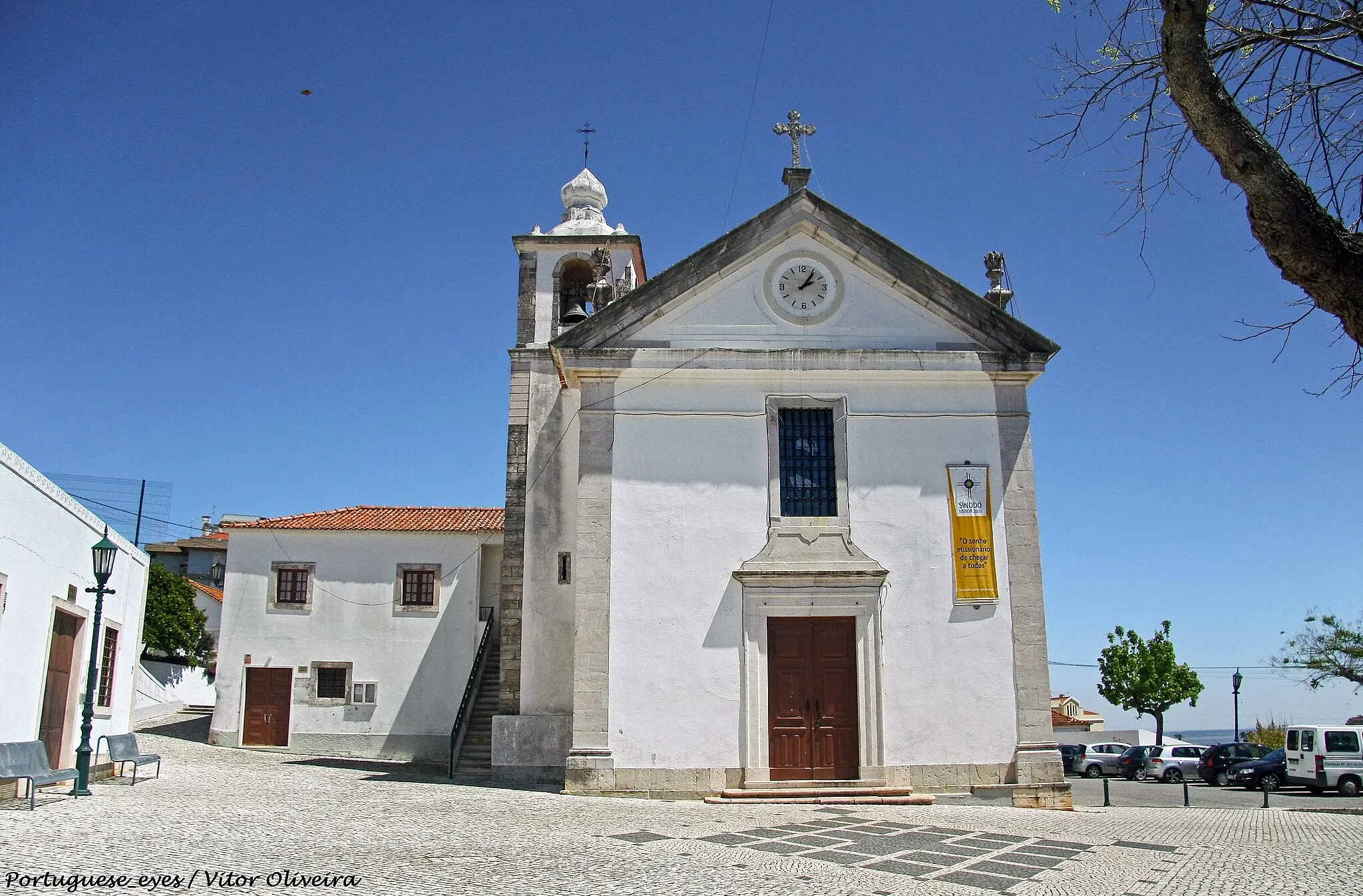 Photo showing: Fundada no século XIV, a igreja paroquial de Vialonga foi objecto de várias campanhas de obras (principalmente no século XVI) que lhe conferiram o seu aspecto actual, um volume compacto, onde impera a ausência de elementos decorativos. Apenas a fachada é animada por um portal rectilíneo e pela janela do coro, sendo rematada por frontão triangular com relógio no tímpano. Do lado da torre sineira, num plano mais recuado, encontram-se as dependências anexas.
Esta austeridade arquitectónica contrasta fortemente com a decoração do interior, que tira partido da conjugação da talha, da pintura e do azulejo, num repositório muito caraterístico da primeira metade do século XVIII. O espaço, de nave única coberto por abóbada de berço, articula-se com a capela-mor, profunda, através de um arco triunfal de volta perfeita, flanqueado por dois altares de talha dourada seiscentista. Para além do púlpito de mármore com balaústres, no corpo do templo abrem-se várias capelas, uma das quais - da Ordem Terceira de São Francisco - com azulejos relatando a vida do santo patrono da Ordem, de cerca de 1735 e atribuídos aos pintores Bartolomeu Antunes e Nicolau de Freitas, que muitas vezes trabalharam em conjunto (SIMÕES, 1979, p. 337). Por sua vez, a capela baptismal, à entrada, conserva o retábulo quinhentista (1535), de mármore (AZEVEDO, FERRÃO, GUSMÃO, 1963, p. 86). Ainda na nave, as paredes são revestidas por painéis de azulejo, com 21 cenas da Vida da Virgem, executados, muito possivelmente, entre 1745-50 (SIMÕES, 1979, p. 337). As cartelas inferiores exibem figurações dos símbolos marianos das litanias.
Na capela-mor, a talha e o azulejo articulam-se, preenchendo a totalidade da superfície parietal, uma vez que, no primeiro registo, se encontram painéis cerâmicos com cercaduras de folhagens e figurações de anjinhos a brincar (c. 1710) (SIMÕES, 1979, p. 337). Sobre este silhar, o revestimento de talha dourada enquadra pinturas e nichos com imaginária. O retábulo-mor, também de talha dourada seiscentista, apresenta tribuna coberta por uma pintura representando a Assunção da Virgem.
Uma última referência para os azulejos da sacristia, com figurações de episódios profanos, com músicos e jardins, que remontam a cerca de 1730.
Através das datações dos azulejos, e de alguma talha, é possível acompanhar as diferentes fases ou campanhas decorativas de que a igreja foi objecto e que se prolongaram por toda a primeira metade do século XVIII. O primeiro espaço a ser intervencionado foi a capela-mor, sendo também aquele que exprime, de forma mais evidente, a ideia de igreja forrada a ouro. De facto, na liturgia barroca, o altar-mor era o centro para o qual convergiam todos os percursos do templo, e onde se expunha o Santíssimo Sacramento, o que justifica a primazia deste espaço na actualização estética, e também litúrgica.
Por outro lado, e em conformidade com a invocação da igreja, a iconografia incide, maioritariamente, sobre a vida de Nossa Senhora, intercessora privilegiada e exemplo de conduta para os fiéis, numa mensagem directa, complementada pela tela da tribuna, evocativa da Assunção da Virgem.
Nesta medida, a mensagem teológica era eficazmente transmitida, integrando-se num espaço profundamente alterado pela presença da pintura, do azulejo e da talha que contribuiam fortemente, com os seus brilhos e cores, para a desmaterialização das paredes e a animação de uma estrutura arquitectónica de grande rigidez, solução muito comum no nosso país durante o período barroco.
(Rosário Carvalho)

www.patrimoniocultural.pt/pt/patrimonio/patrimonio-imovel...