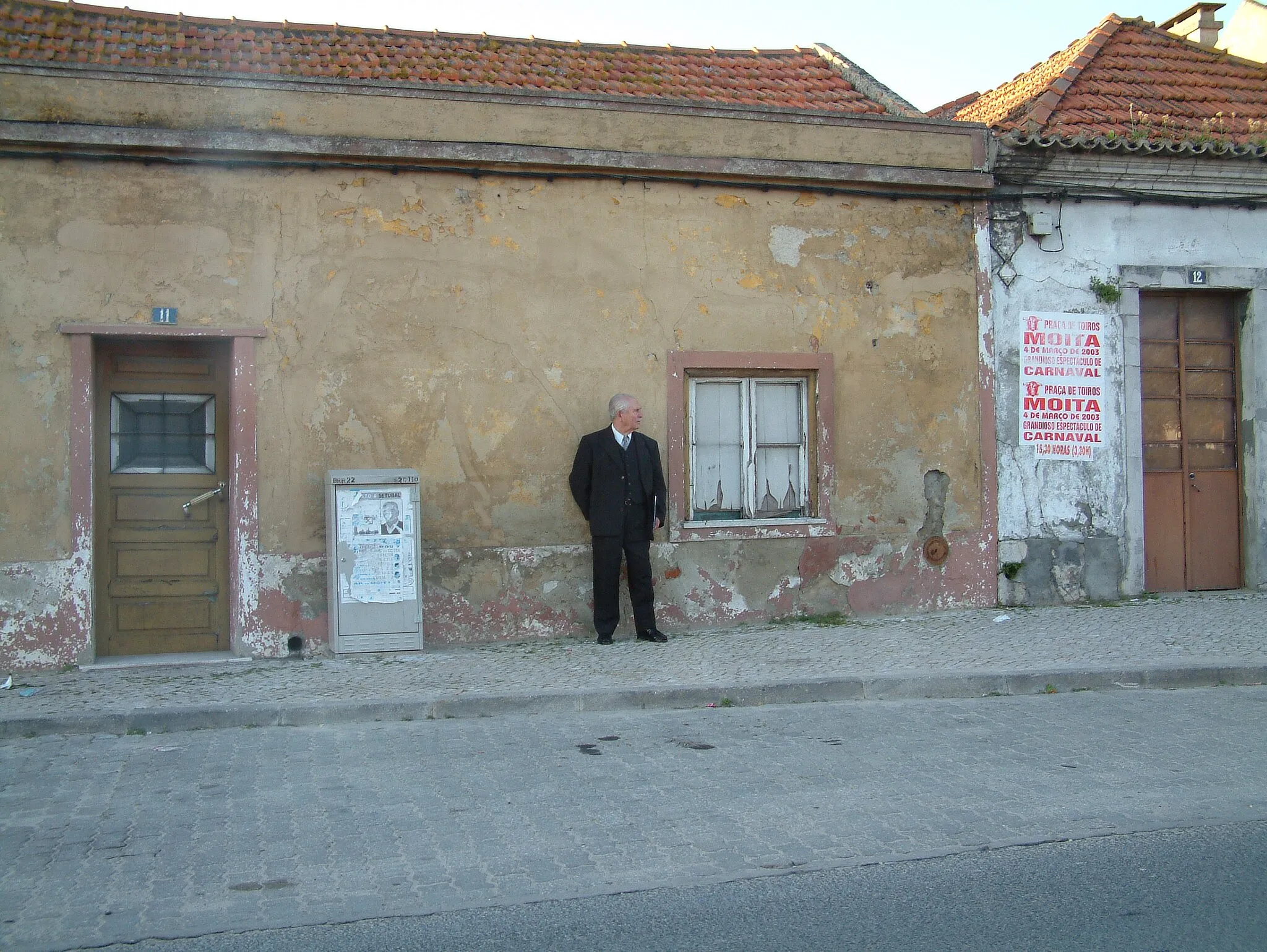 Photo showing: Notice the contrast between the decrepit paint on the walls and the impeccable dress of the old man. Taken in small village "Santo António da Charneca" near Barreiro in Portugal.