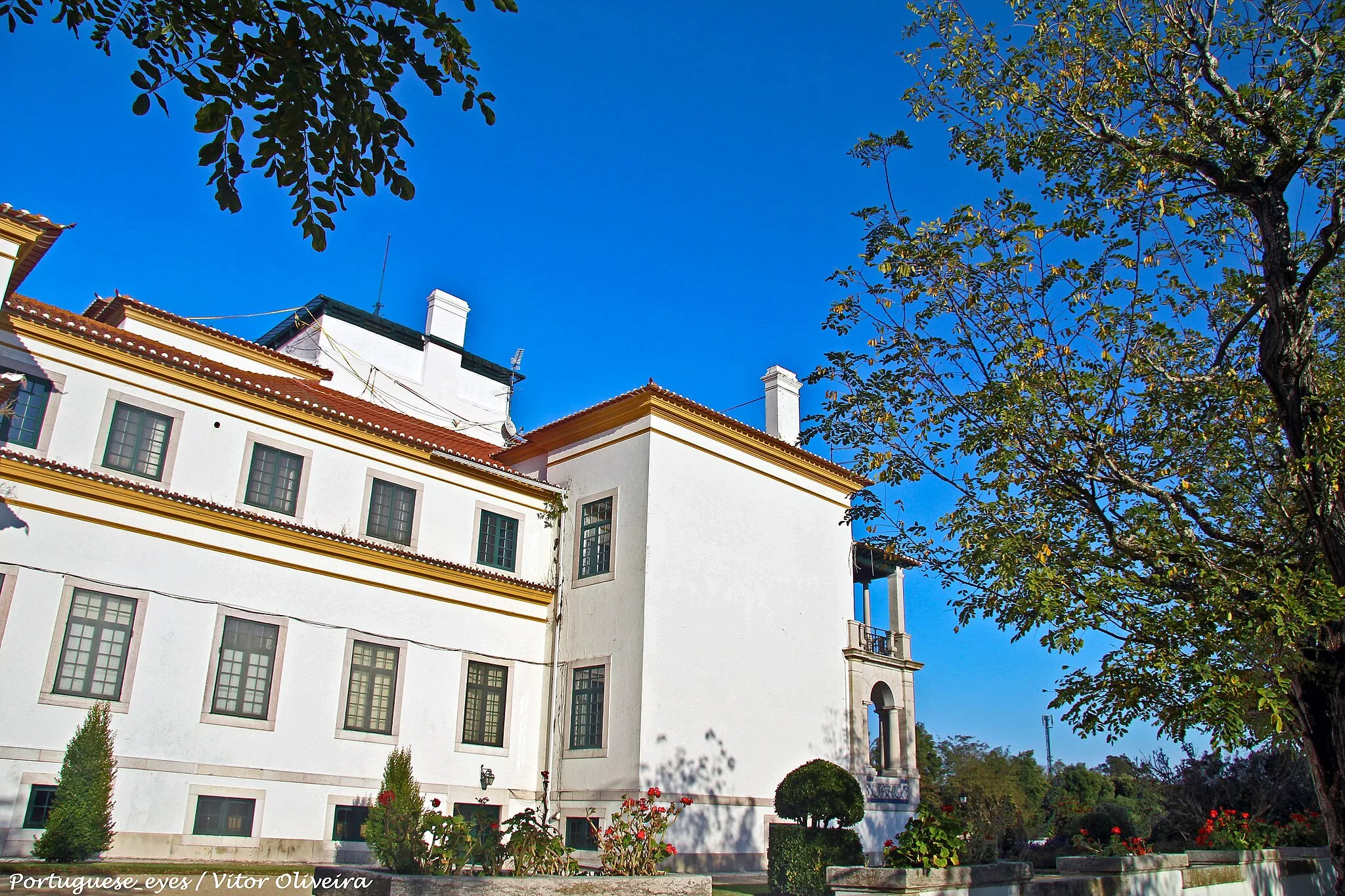 Photo showing: Entre o Montijo e Alcochete, não muito longe das margens do Tejo, o silêncio magnífico envolve o Palácio de Rio Frio e os campos dourados ao fim da tarde. Já lá vão os tempos em que este palácio era a casa abastada de uma das maiores herdades do país. Em 1892, ainda antes de o palácio existir, Rio Frio era uma propriedade de 17 mil hectares, onde o veterinário José Maria dos Santos mandou plantar uma gigantesca vinha, o que significa qualquer coisa como dez milhões de cepas, constituindo, para a época, uma exploração vinícola sem rival no mundo.
Nas gerações seguintes, a herdade continuava a ser uma das maiores do pais e foi deixada em herança a um sobrinho de José Maria dos Santos, Alfredo Santos Jorge, que mandou construir o Palácio de Rio Frio no princípio do século. Não se sabe quem foi o arquitecto desta casa por terem desaparecido num incêndio todos os arquivos que o identificariam, mas os azulejos que decoram o exterior e o interior da casa foram encomendados a Jorge Colaço. Maria de Lourdes Pereira Lupi d'Orey, actual proprietária do Palácio de Rio Frio e sobrinha -neta de Alfredo Santos Jorge, ainda se lembra da magia da vida na herdade, quando para aqui veio viver, em criança, com os pais. Era uma época em que o tempo passava devagar, ao ritmo das estações e das épocas agrícolas. Produziam-se na herdade todos os produtos básicos e necessários à vida diária da família: pão, manteiga, farinha, ovos, leite, arroz, legumes, carne de gado, etc. Eram uma família grande, profundamente ligada ao campo e à lavoura, e, dos vários irmãos de Maria de Lourdes, foi José Samuel Lupi que se destacou como figura tauromáquica de relevo no seu tempo.
Apesar da proximidade de Lisboa, uma estadia no Palácio de Rio Frio tem todos os ingredientes para um delicioso mergulho na natureza e permite usufruir de todos os prazeres do campo, num ambiente simpático e acolhedor.

RNET n.º 4510 - Turismo de Habitação www.center.pt/PT/detalhes-palacio-de-rio-frio