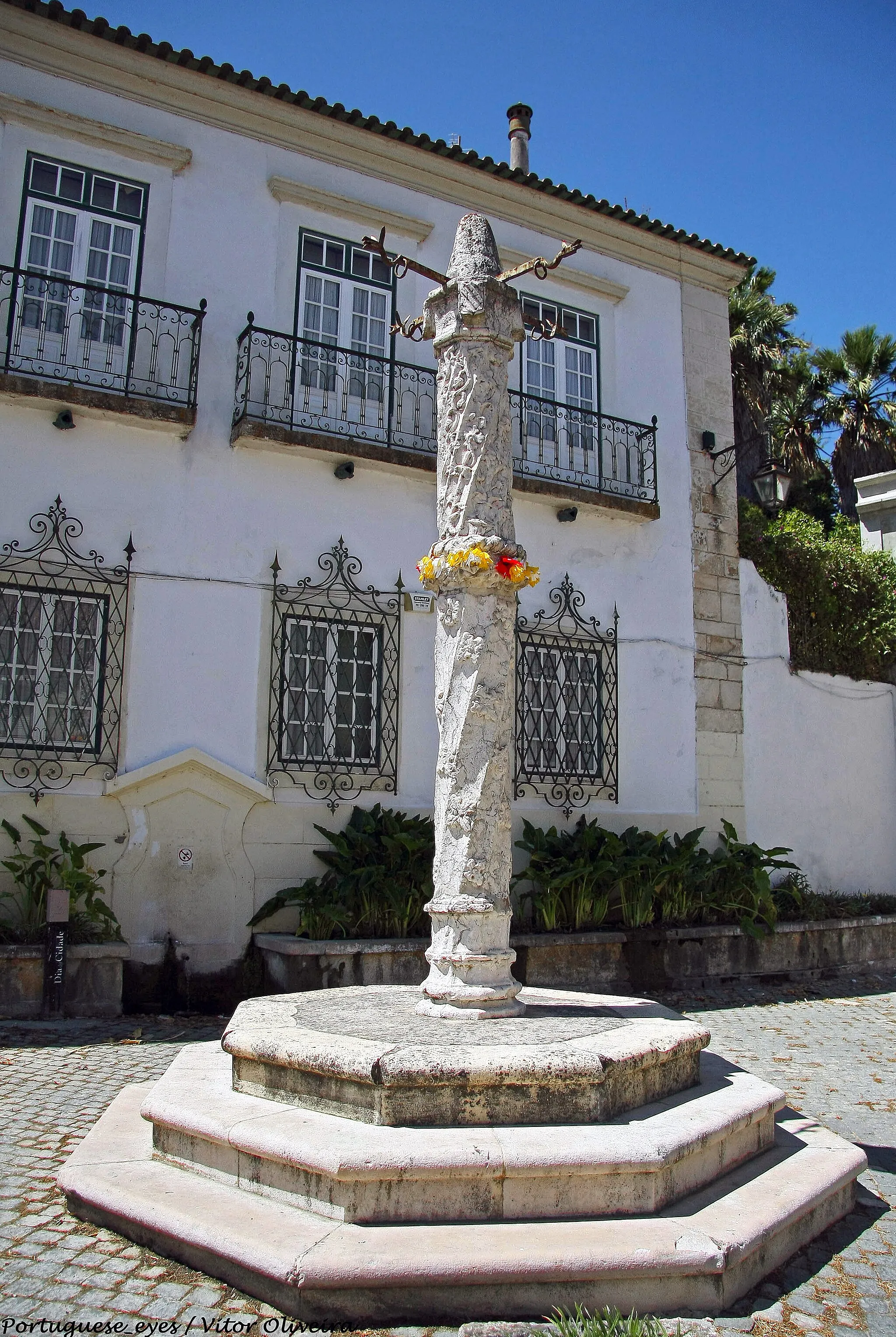 Photo showing: Povos, no actual concelho de Vila Franca de Xira, teve foral dado por D Sancho I em 1195, atestando este facto a importância que o pequeno burgo e o seu castelo, erguido no alto do monte do Senhor da Boa Morte e sobranceiro ao Tejo, detinha então em terrenos de Reconquista. Este primeiro foral, confirmado em 1218 por D. Afonso III, foi dado de novo por D. Manuel, em 1510, a par de algumas vilas limítrofes (caso de Castanheira e Vila Franca). O pelourinho que hoje se ergue diante da antiga Casa da Câmara, no Largo da Forca, datará dos anos subsequentes, sendo de clara tipologia manuelina. Note-se, a este respeito, que a designação do largo (então o terreiro central da povoação) poderia estar relacionada com a existência de uma forca permanente neste local, mas não deve respeitar ao pelourinho em si, visto que estas construções não eram utilizadas para a execução da pena terminal. 
O pelourinho de Povos é uma peça muito semelhante aos vizinhos pelourinhos de Vila Franca de Xira e Alverca do Ribatejo, embora estes sejam reconstruídos (a partir de fragmentos originais). Sobre um soco composto por três degraus octogonais levanta-se a pequena base facetada onde assenta o fuste, formado por dois blocos cilíndricos torsos, ligados por anel duplo de torcidos e folhagem. O primeiro bloco do fuste, ligeiramente mais comprido, é decorado com rosetas entre as estrias helicoidais, e o segundo é decorado com motivos vegetalistas e grotescos. O capitel, em tronco cilíndrico, exibe três pedras de armas dos Ataídes, senhores de Povos, Castanheira e Cheleiros, por causa das quais se tem considerado ser o pelourinho uma encomenda de D. António de Ataíde, 1º Conde da Castanheira (com título outorgado em 1521). No mesmo capitel, e entre os brasões, uma cabeça em relevo lembra ainda o capitel da picota de Alverca do Ribatejo, onde cabeças de anjos se alternam com escudos de Portugal. Por fim, temos um remate cónico de topo desbastado, que conserva ainda os quatro ferros dispostos em cruz, com argolas nas extremidades, fixos entre o capitel e o remate. Quando a este, poderá não ser uma peça original, já que se encontram referências a uma pinha elipsoidal (MALAFAIA, Ataíde, 1997).

É provável que a estrutura tenha sofrido alguma intervenção não documentada depois de 1850, quando é referida a existência de quatro degraus, em vez dos três que actualmente se vêem (AMARAL, João José, 1997); é Virgílio Correia quem dá notícia, em 1924, de um restauro feito a expensas de uma família da terra (CORREIA, Vergílio, 1924). Nessa data, o pelourinho tinha os degraus embebidos no pavimento, ficando apenas o degrau cimeiro à vista. Mais tarde, no início dos anos 70, o pelourinho necessitou de escoramento, e chegou a ser desmontado para restauro. É possível que a moderna nivelação do largo tenha sido realizada na mesma altura, revelando três degraus da plataforma. SML www.patrimoniocultural.gov.pt/pt/patrimonio/patrimonio-im...