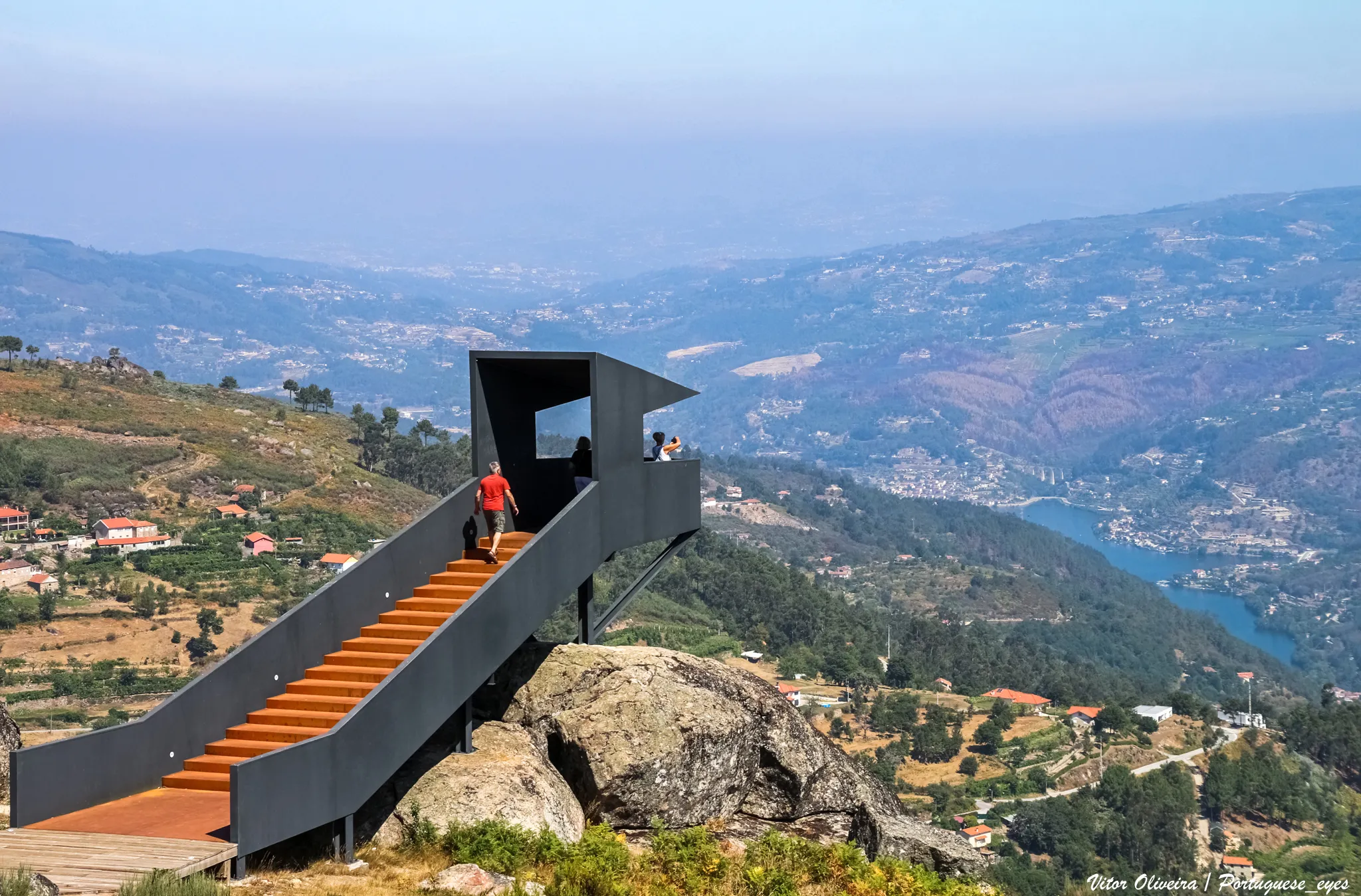 Photo showing: Situado em plena Serra do Montemuro, o projeto contempla uma paisagem caracteristicamente composta por mosaicos, tais como aglomerados rurais, agricultura e floresta, capazes de albergar uma diversidade faunística e florística imensa. Mais concretamente localiza-se no espaço adjacente à Rua do Miradouro, na localidade de Marcelim, pertencente ao concelho de Cinfães, num troço próximo da Capela de Nossa Senhora dos Milagres. O projeto consiste na construção de dois pontos de observação estrategicamente colocados, um no patamar superior, com vistas orientadas para o vale do rio Bestança, e outro no patamar inferior, com vistas orientadas para a foz do rio Bestança e o vale do rio Douro. A presente operação visa atribuir uma nova funcionalidade ao espaço como espaço público de lazer, tirando o melhor proveito das mais-valias e vistas que esta área proporciona, tendo em conta as suas características naturais, de compatibilização com as mesmas e numa perspetiva de sustentabilidade, indo assim ao encontro dos objetivos do eixo prioritário de investimento, que se baseiam na conservação, proteção, promoção e desenvolvimento do património natural e cultural. Com a implementação da intervenção proposta, o município de Cinfães pretende alavancar o investimento no Turismo de Natureza, sendo este considerado como um dos principais produtos turísticos prioritários da Região do Norte, como emanado na Agenda Regional do Turismo. O projeto tem como objetivo a criação de um espaço de visita e lazer, permitindo a valorização dos recursos naturais existentes no Município, com equipamentos e infraestruturas de apoio ao seu funcionamento, nomeadamente, pavimentações, zona de estacionamento, espaço pedonal e zonas com mobiliário urbano. É também pretendida a criação de plataformas, tipo observatórios da natureza, para que os visitantes possam usufruir da paisagem do local. A operação visa também a aposta num turismo acessível para todos, promovendo a acessibilidade a pessoas com deficiência ou incapacidade. Neste sentido o projeto prevê a colocação de um elevador para pessoas com mobilidade reduzida, tornando possível o acesso da parte superior por pessoas em cadeira de rodas ou com dificuldade de locomoção. A proposta tem como premissa a requalificação do espaço a intervencionar, tornando-o um local de usufruto público dedicado ao lazer e à fruição da paisagem, tendo como base a incomparável potencialidade das vistas que esta zona proporciona, permitindo assim alcançar um objetivo primordial que se traduz no aumento do número de visitantes a sítios de património cultural e natural. Pretende-se que o Observatório da Natureza de Marcelim constitua uma mais-valia como espaço público para todo o concelho e a região Norte do país, constituindo-se como um espaço utilizável por todos os visitantes e tornando-se num ponto atrativo para o Turismo de Natureza. Desta forma, o município visa promover o equilíbrio necessário entre o desenvolvimento, o investimento e o respeito pela biodiversidade e conservação da natureza, adotando e concretizando políticas e ações que visam o Turismo Sustentável. cm-cinfaes.pt/acao-municipal/portugal-2020-mm/item/3662-o...