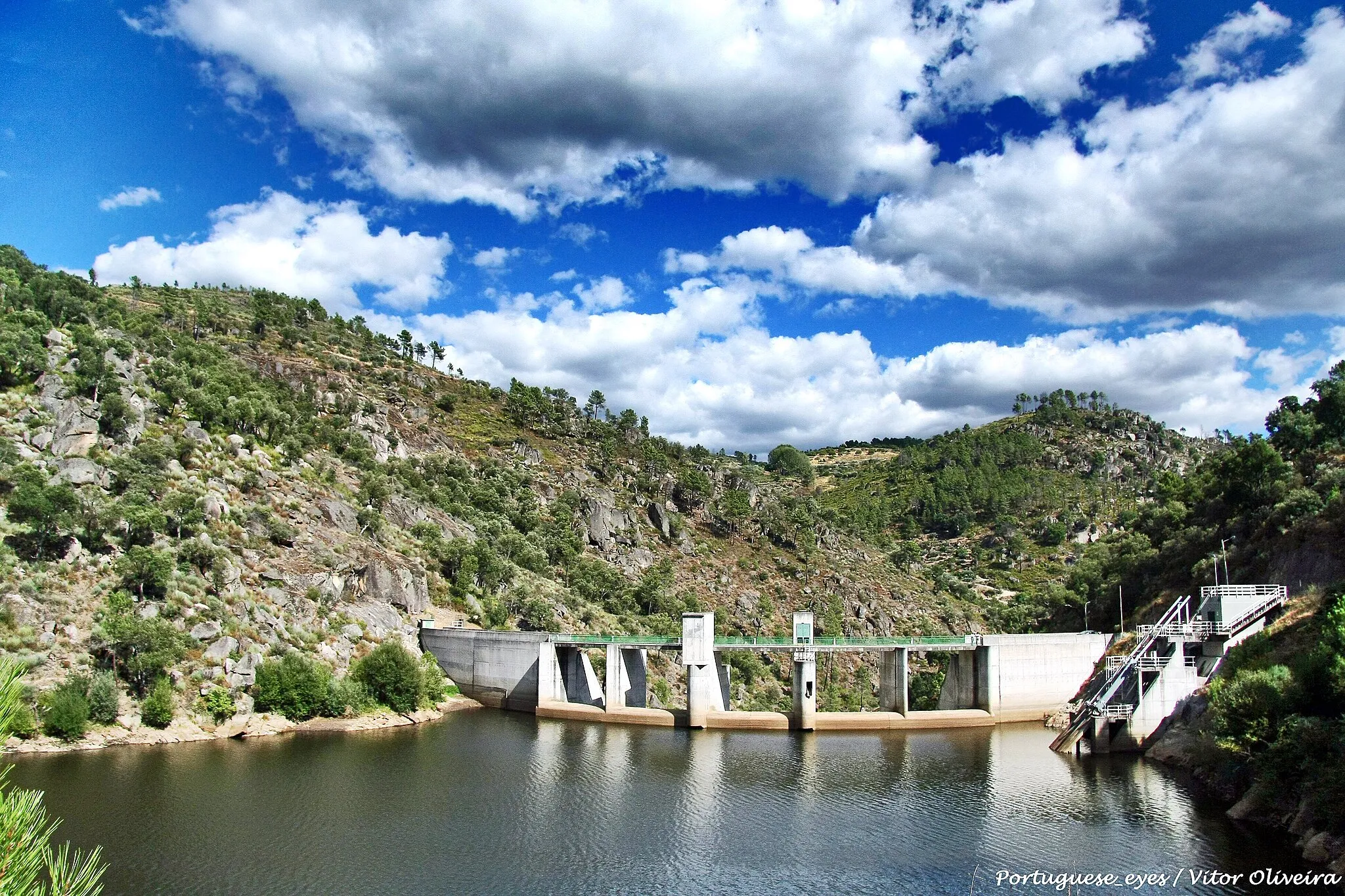 Photo showing: A barragem de Rebordelo localiza-se no concelho de Vinhais, distrito de Bragança, Portugal. Situa-se no rio Rabaçal. A barragem foi projectada em 1999 e entrou em funcionamento em 2004.
Barragem
É uma barragem de arco e gravidade em betão. Possui uma altura de 35,5 m acima da fundação e um comprimento de coroamento de 127 m. O volume de betão é de 19.000 m³. Possui uma capacidade de descarga máxima de 1.480 m³/s.
Albufeira
A albufeira da barragem apresenta uma superfície inundável ao NPA (Nível Pleno de Armazenamento) de 46 hectares e tem uma capacidade total de 3,13 hm³. As cotas de água na albufeira são: NPA de 380 metros, NMC (Nível Máximo de Cheia) de 386 metros e NME (Nível Mínimo de Exploração) de 378,5 metros.
Central hidroeléctrica

A central hidroeléctrica é constituída por um grupo Kaplan com uma potência instalada de 8,75 MW. A energia produzida em média por ano é de 24 GWh. pt.wikipedia.org/wiki/Barragem_de_Rebordelo