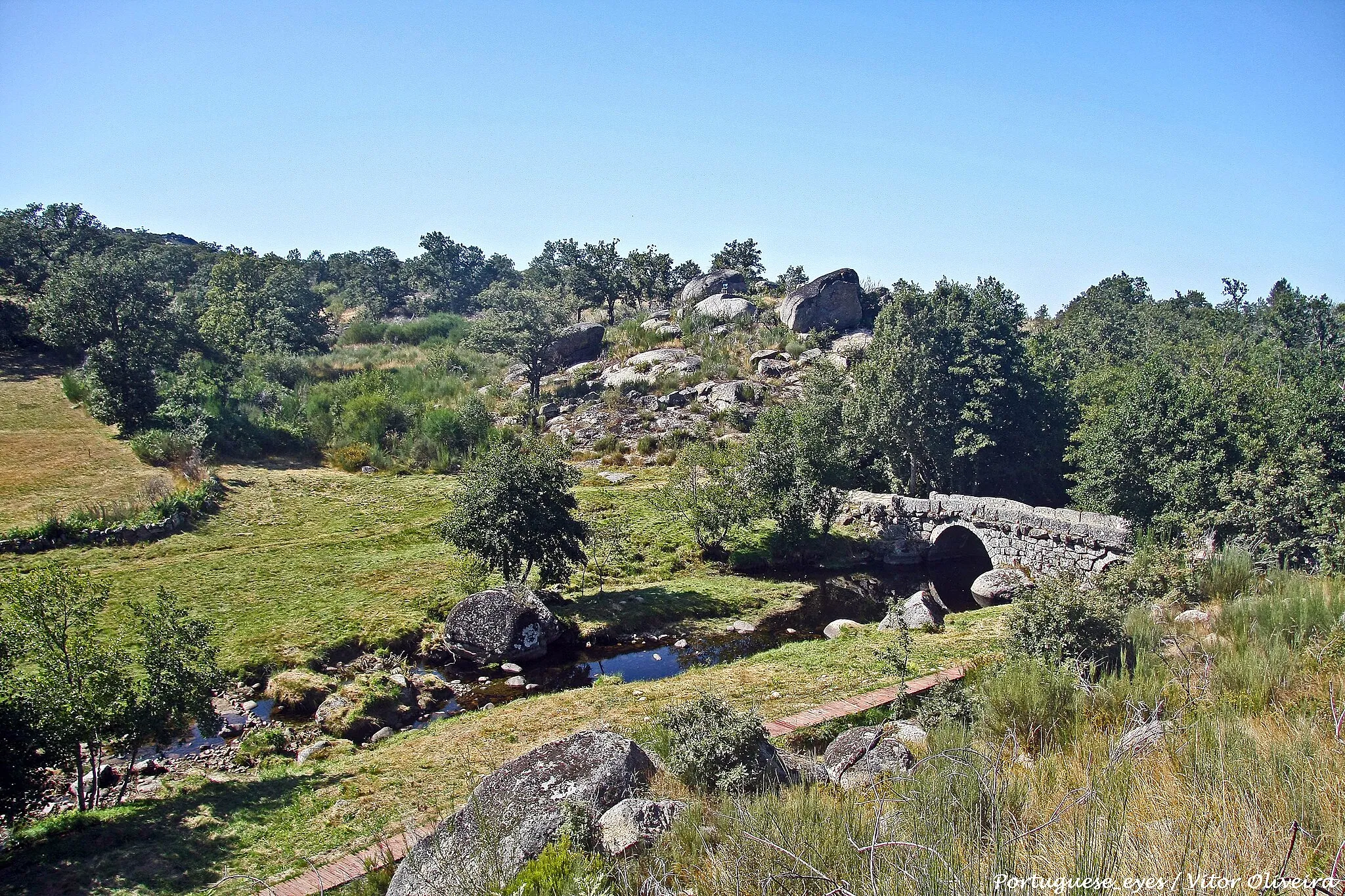 Photo showing: A Ponte da Panchorra, classificada como Monumento de Interesse Público desde 2013, integra o percurso turístico-cultural da Rota do Românico. Implantada a cerca de 1000 metros de altitude, unindo as margens do rio Cabrum, a Ponte da Panchorra é um belíssimo exemplo de arquitetura vernacular [tradicional]. Ponte de dois arcos, apresenta aparelho regular nas aduelas [pedras que formam o arco] e irregular na silharia [pedras] da restante estrutura, o que pode indicar um trabalho de mestres locais ou regionais, destinado a suprir as necessidades de acesso da comunidade às suas propriedades agrícolas e silvícolas. Nesse sentido, distancia-se em importância e técnica das suas congéneres, edificadas a jusante, nomeadamente as pontes de Ovadas, Lagariça e Nova, quase na foz do Cabrum. Não deixa, porém, de ser um exemplo de infraestrutura comunitária. A travessia aproveita os afloramentos das margens do rio para apoiar os seus pilares, sobre os quais assenta o tabuleiro horizontal com guardas, conferindo-lhe a robustez necessária à passagem de carros agrícolas e à circulação de gado. Embora a Panchorra seja referida já nas Inquirições[inquérito administrativo] de 1258, só no século XVI se separou do termo de Ovadas, onde se situava o antigo centro religioso da freguesia medieval. Tornou-se, então, curato [paróquia], sendo a ermida de São Lourenço o novo polo religioso. www.guiadacidade.pt/pt/poi-ponte-da-panchorra-285521