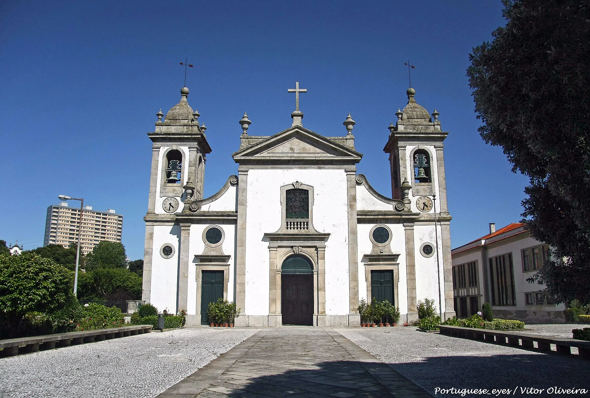 Photo showing: Esta igreja foi construída nos princípios e a par da fundação de Portugal e era conhecida, pelo nome de “Igreja de S. Miguel de Moroça”. Construção antiga, pequena e baixa, revestida de pedra, por dentro e por fora. Traçado um tanto semelhante à velha Igreja de Cedofeita, ou qualquer outra românica. Nada existe nesta freguesia que “pertença” a construções românicas, portanto existe a hipótese de ter sido demolida e sobre ela se ter edificado a actual, entre 1568 a 1578.
É de realçar nesta edificação o retábulo da capela-mor de talha dourada com colunas salomónicas e decoração de parras, cachos de uvas, pombas, folhas de acanto e cabeças aladas, característica do séc. XVIII. Este retábulo foi concluído em 1714 e dourado pelo pintor portuense Manuel Ribeiro. O remate, em estilo rocaílle, deverá datar de 1762 e 1778.
Sobre o guarda-vento existe o coro que em tempos teve arcos e abóbada, sofrendo obras de modificação em 1873, a fim de dar passagem da torre Sul para a Norte, uma vez que esta não pode ter escadas de acesso devido aos dois grandes pesos graníticos das respectivas cordas do relógio. Interiormente encontramos um templo de três naves, com cinco arcos de cada lado, sobre quatro colunas em cantaria com base e capitel e mais duas embebidas no frontispício, em cantaria à vista. O chão, talhado a granito, em sepulturas, outrora muradas, e onde jazem as cinzas de muitos dos nossos antepassados; com tampos em macacauba e pau cetim.
O tecto das naves, em castanho velho encerado, com ornatos em talha dourada. Tem setenta e cinco caixotões na nave central e quarenta e cinco em cada uma das laterais. Estes, caixotões foram colocados em 1735 e copiados pelos então existentes no tecto da capela-mor. A iluminação natural faz-se por janelas rasgadas nas paredes, das quais por obra mandada fazer a expensas de D. Adelina Leite Nogueira Pinto Domingos de Oliveira, foram retiradas as grossas grades de ferro e substituídas por singelos vitrais artísticos.
Neste percurso em direcção ao altar-mor, associámo-lo à caminhada ou à preparação em direcção à zona principal do culto encontrámos, diversos altares:
À nossa direita, o Retábulo do Senhor Morto, apresenta a curiosidade de a mesa do altar ser um mostrador rectangular de linhas simples, onde jaz a belíssima escultura articulada do Senhor Morto (século XIX) que era exposta no passo do Calvário ao terminar a Procissão dos Passos. O retábulo tem ao centro uma imagem da Senhora do Rosário e aos lados as de São José e a de S. João Baptista. Duas esculturas do século XVII, provenientes da Capela de Sant’Ana.
Do lado esquerdo o Retábulo das Almas ou da Senhora do Carmo, bastante trabalhado nas colunas e entablamentos, é uma obra do século XIX com características maneiristas.
A este retábulo preside uma belíssima escultura do século XX, da autoria de José Ferreira Tedim.
Nas laterais estão as esculturas de São Bento e São Brás. Mais à frente, do lado direito o Retábulo do Senhor dos Passos. Preside a este retábulo a realística imagem do Senhor dos Passos que sai na majestosa procissão que anualmente percorre as ruas de Leça da Palmeira debaixo de elevado respeito e devoção.
Trata-se de uma imagem com o corpo bem modelado, não é uma imagem de roca. Em mísulas laterais estão duas imagens, o Senhor preso a coluna e “Ecce Homo”. Do lado oposto, o Retábulo do Coração de Jesus, na capela que actualmente lhe é dedicada, mas outrora já o foi a S. José, a Santa Quitéria e também a S. Lourenço.
Salienta-se deste retábulo ensamblado em 1906, mas sendo uma peça do barroco a escultura do Santíssimo Coração de Jesus, em tamanho natural, da autoria do mestre Francisco Couceiro, e oferecida por D. Adelaide Leite Nogueira Pinto.
Nos nichos laterais a Rainha Santa Isabel feita por Teixeira Lopes e o Beato Nuno Álvares Pereira. Lateralmente ao frontispício da capela-mor temos do lado esquerdo o Retábulo de São Miguel, onde encontramos a imagem do padroeiro da freguesia numa escultura que constitui uma verdadeira obra prima do mestre Guilherme Tedim, encomendada pela respectiva confraria em 1933. No mesmo altar, lateralmente temos Santo António e São João de Brito.
Do lado direito o Retábulo de Nossa Senhora de Fátima, onde ao centro temos a imagem da Senhora de Fátima esculpida por mestre Guilherme Tedim apresenta uma candura e transmite uma bondade das suas feições faciais que tocam o coração de quem a observa. No mesmo altar temos Santa Lúzia, advogada dos olhos, e o Mártir São Sebastião oriundo da respectiva capela que ficava do outro lado da rua. Já sobre o altar duas imagens, mais recentes, dos Beatos Francisco e Jacinta.
Prosseguindo o nosso caminho chegamos à Capela-Mor, com o seu retábulo, muito antigo, construído em madeira e profusa talha dourada. Ao centro deste retábulo um trono maravilhoso, muito bem concebido e executado, lateralmente temos as imagens de Santa Apolónia, advogada das dores de dentes, Santiago e São João Evangelista oriundo da Capela do Corpo Santo. Esta Capela-Mor tem lambrim em azulejo. À entrada da Capela-Mor está a imagem da Senhora da Conceição, proveniente do Convento Franciscano da Quinta da Conceição e que será oportunamente objecto de uma referência própria.
Na sacristia destacamos, numa moldura a “autêntica” (certidão da relíquia) do Santo Lenho e a respectiva cruz-relicário que costuma ser levada sob o palio nas Procissões dos Passos do Senhor.
À saída o baptistério fechado por umas grades em ferro forjado com um interessante e pormenorizado trabalho. No seu interior para além da Pia Baptismal, uma imagem de Santa Isabel, prima de Santíssima Virgem. Vê-se assim que vale a pena entrar e observar a nossa igreja com atenção, por isso aproveitem porque se trata de uma das poucas igrejas frequentemente abertas.

(Texto: Eng.º Rocha dos Santos) www.leca-palmeira.com/igreja-matriz-de-leca-palmeira/