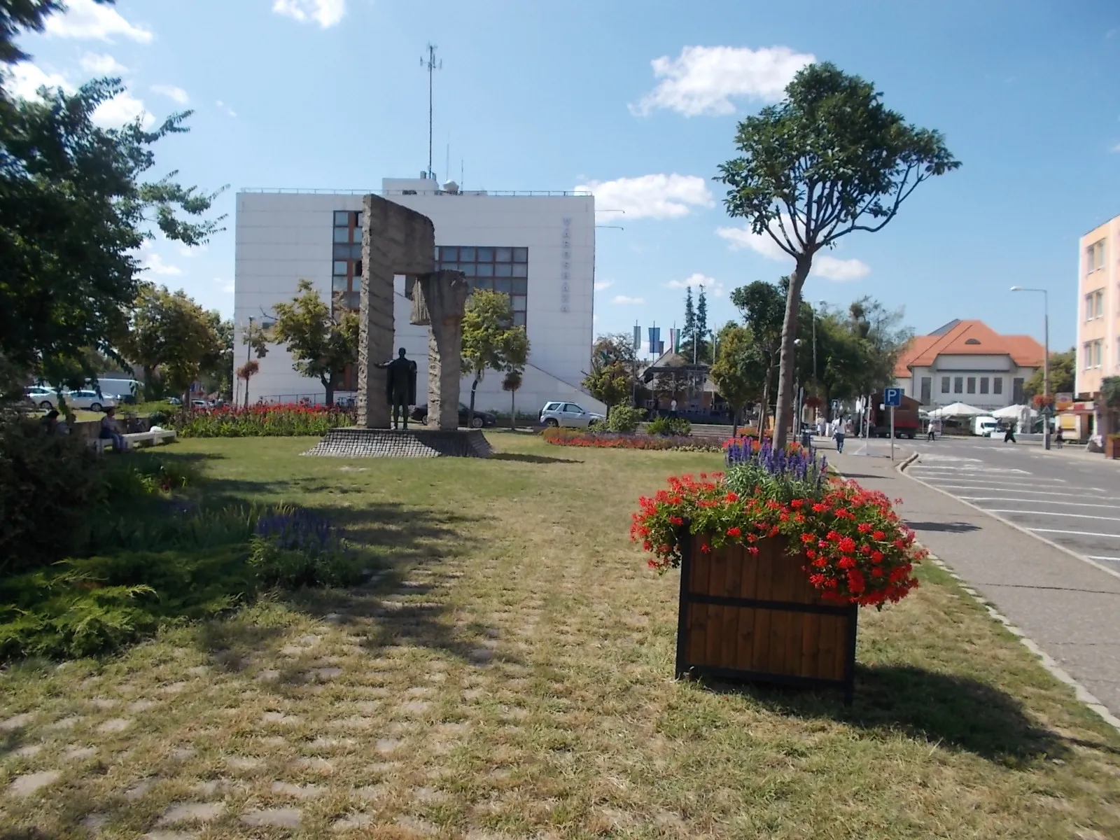 Photo showing: Town Hall (at back) and Freedom memorial by István Kiss, (1981, concrete gate is 8 meters high and 3 meters wide, two and a half meters high male with flowers in his hand stand on a basalt cube pile, a replacement of the WWII memorial?, planned in 1975, a simple agricultural worker figure who enjoying the Life, and the liberation) - Hősök Square, Mátészalka, Szabolcs-Szatmár-Bereg County, Hungary.