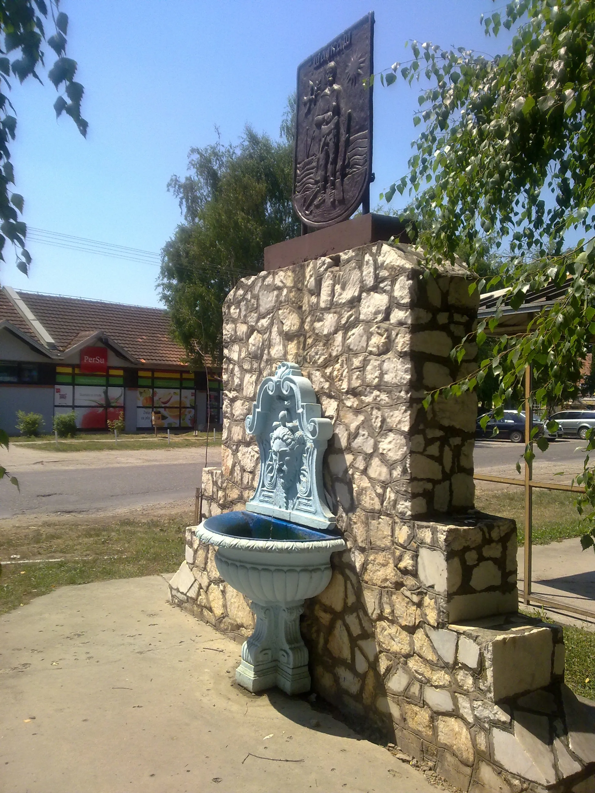 Photo showing: The photo shows the fountain in the centre of the village of Šajkaš (Serbia). Above the fountain, the coat of arms of Šajkaš can be seen.