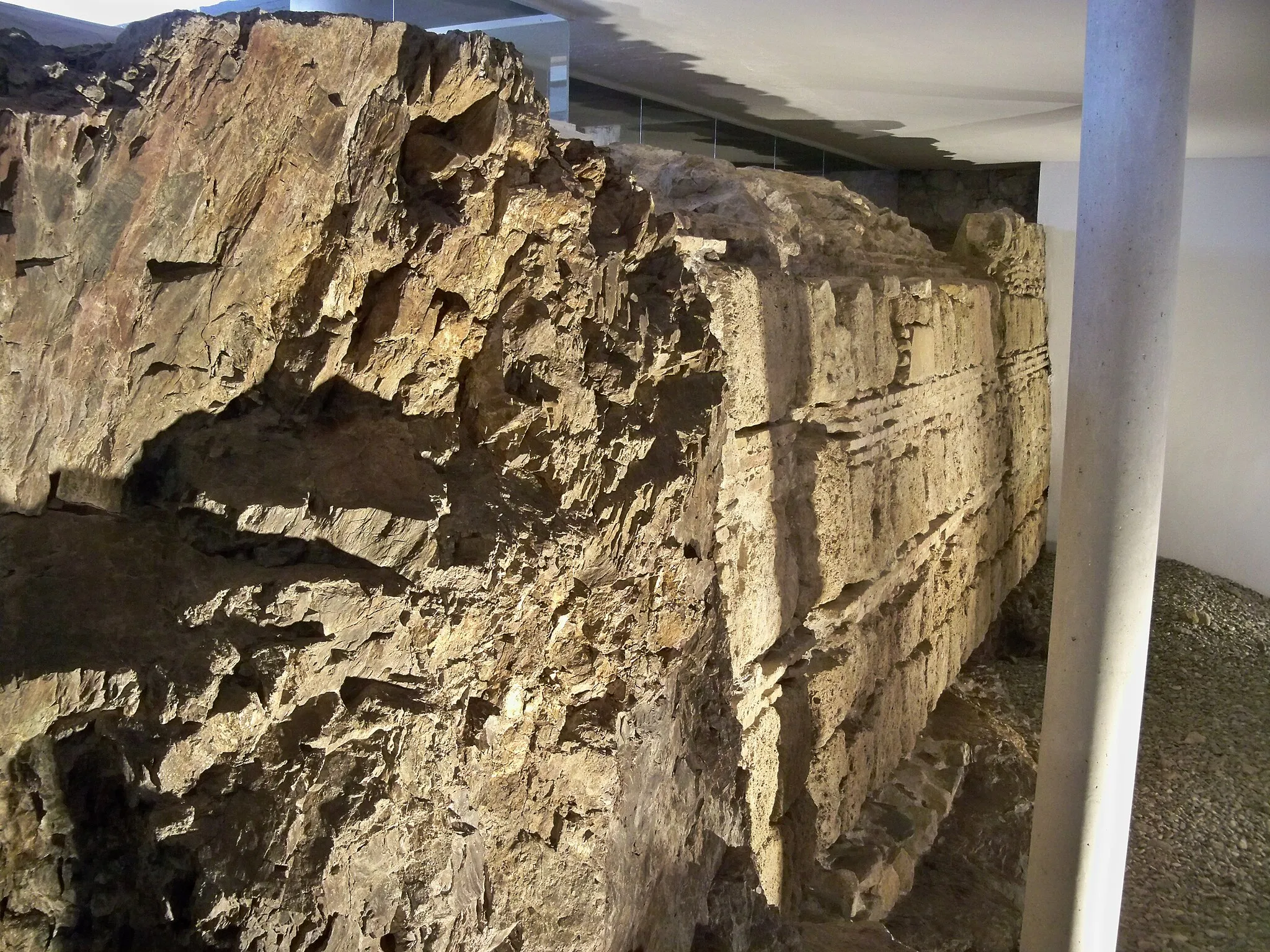 Photo showing: Restos de la antigua muralla medieval de Málaga bajo el edificio del Rectorado de la Universidad de Málaga, en Málaga, España. En el yacimiento arqueológico del Rectorado también se encuentran restos de la muralla de la ciudad fenicia, aunque no están visibles al público. En cuanto a los restos de época medieval, la sección de muralla constituida a base de sillares a soga y tizón pertenece a la época califal (s. XI), mientras que la sección de sillares con verdugadas de ladrillo pertenece a la época almohade (s. XII) y nazarí (s. XIII-XIV). En la parte izquierda de la imagen, de color marrón oscuro, se aprecia lo que fue un antiguo espolón de pizarra que sobresalía de la vertical de la muralla.