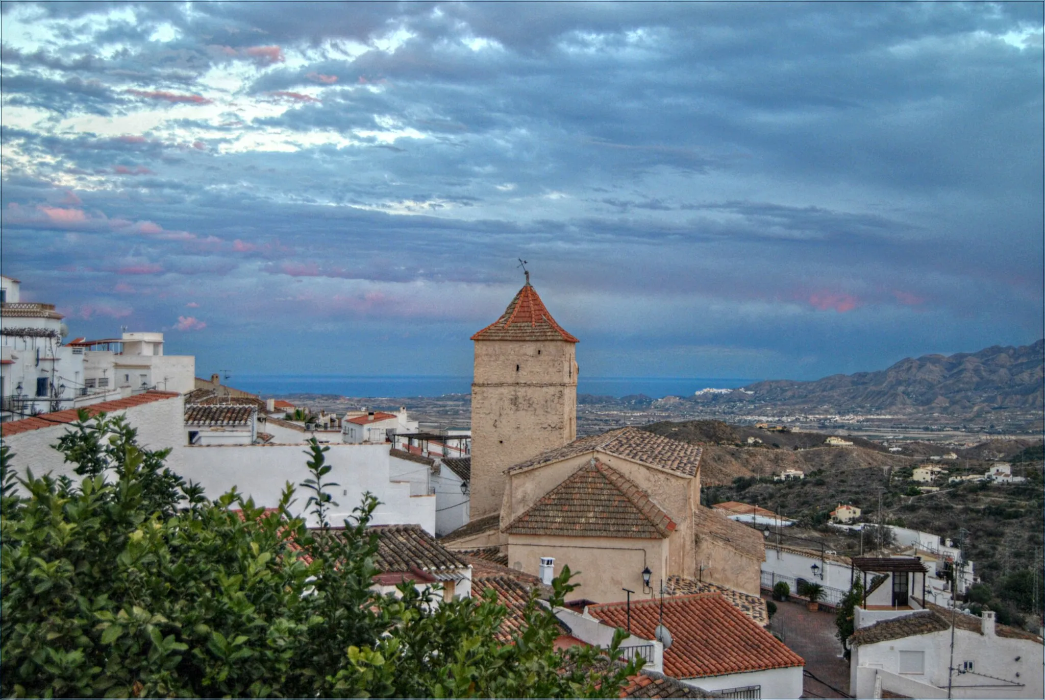 Photo showing: The parish church Iglesia Santa Maria de la Cabeza can be spotted above the village house rooftops, it is located in the Plaza Santa Maria, inside the church there are two oil paintings, one of San Gregorio and the other of the Virgen de la Cabeza, both being the patron saints of the pueblo. Just to the right of the church can be seen the town of Mojacar nestled in the mountains.