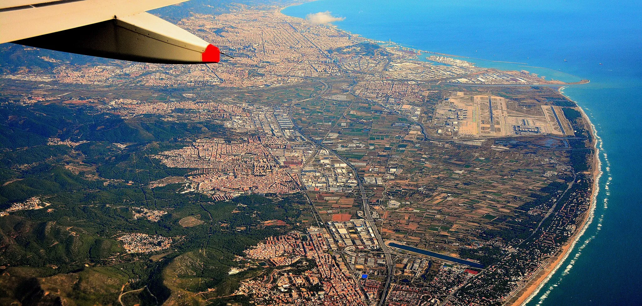 Photo showing: We are looking northeast, from a height of approx 12,000 feet (3650 metres). At top left is part of the city of Barcelona, including its port area. The darker area near the port is the historic hill of Montjuïc, which is believed to be the birthplace of the city.
Centre right is Barcelona's airport, named after the nearest town -  "El Prat". From the airport south-westwards there are many kilometres/miles of popular beaches. Behind the beaches are trees, with houses in between (except near the airport, where there are nature reserves.)

Clearly seen at the bottom of the picture is a long narrow waterway, close to the town of Castelldefels, which was constructed for the 1992 Olympics as a venue for canoe sprint events.