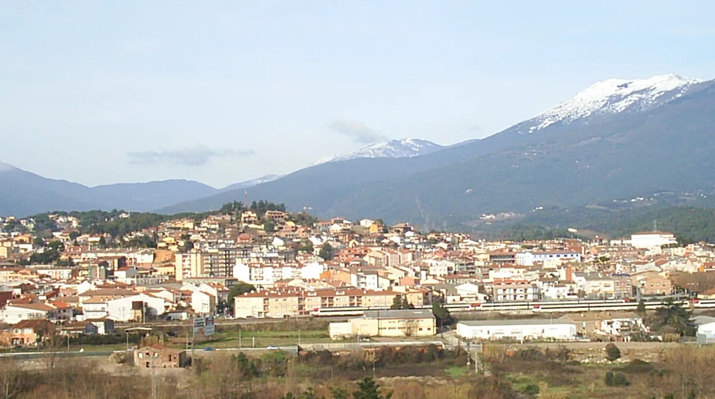 Photo showing: El Turó del Puig de Bellver és el cim en primer terme que està coronat per un campanar. A sota està la ciutat de Sant Celoni i al fons el massís del Montseny