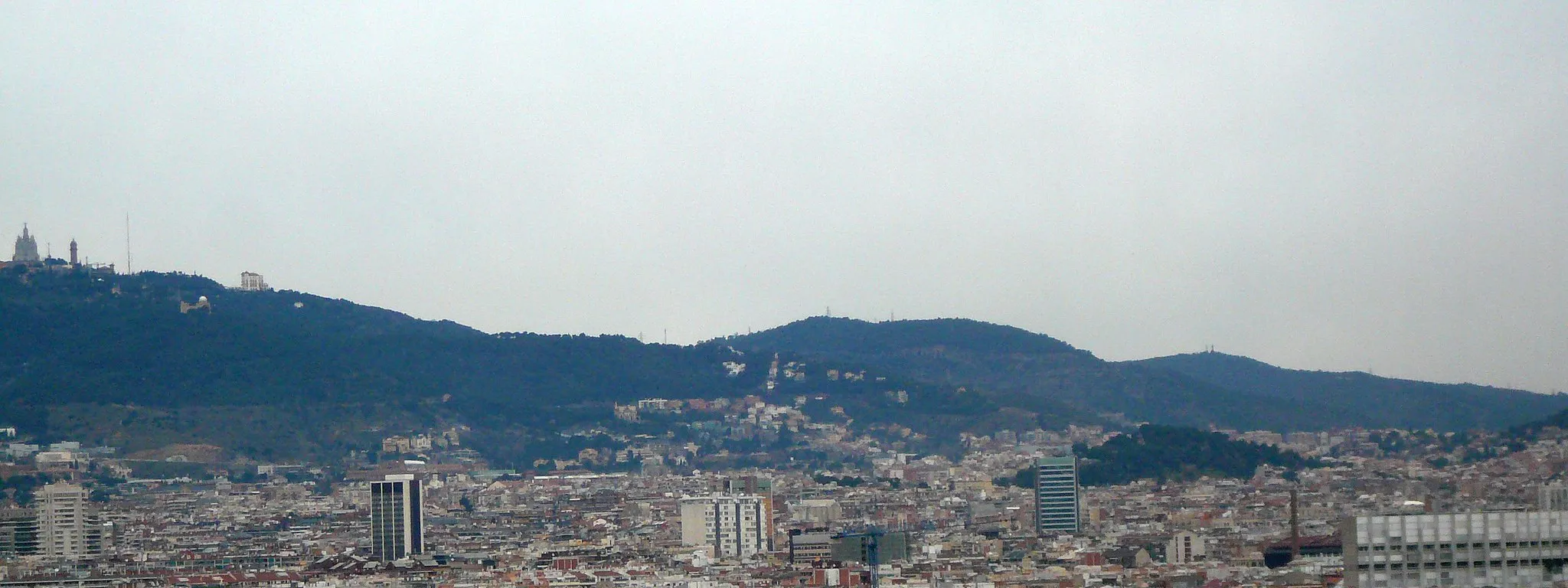 Photo showing: Vista de Collserola des de davant del Palau Nacional de Montjuïc. S'hi veu el Tibidabo (a l'extrem esquerra), amb l'Observatori Fabra al davant, el Turó de Santa Maria (cap al centre de la imatge) i el Turó de Valldaura a la dreta (amb dues construccions al cim, gairebé inapreciables a la imatge: a l'esquerra una antena i a la dreta la torre de vigilància contra incendis).