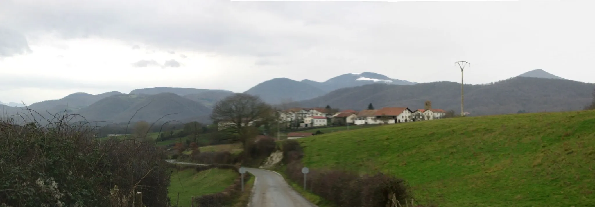 Photo showing: vista general de Illaregui, desde la carretera NA-411 que se acerca al lugar desde el este. A la derecha de descubre la torre de la Iglesia de San Miguel, y a continuación las casas de la calle principal, cubiertas parcialmente por el caserio situado al sur de esa calle; al fondo la cadena montañosa, divisoria de las aguas del Atlántico y del Mediterráneo. La cima más alta es la del Arnaizu