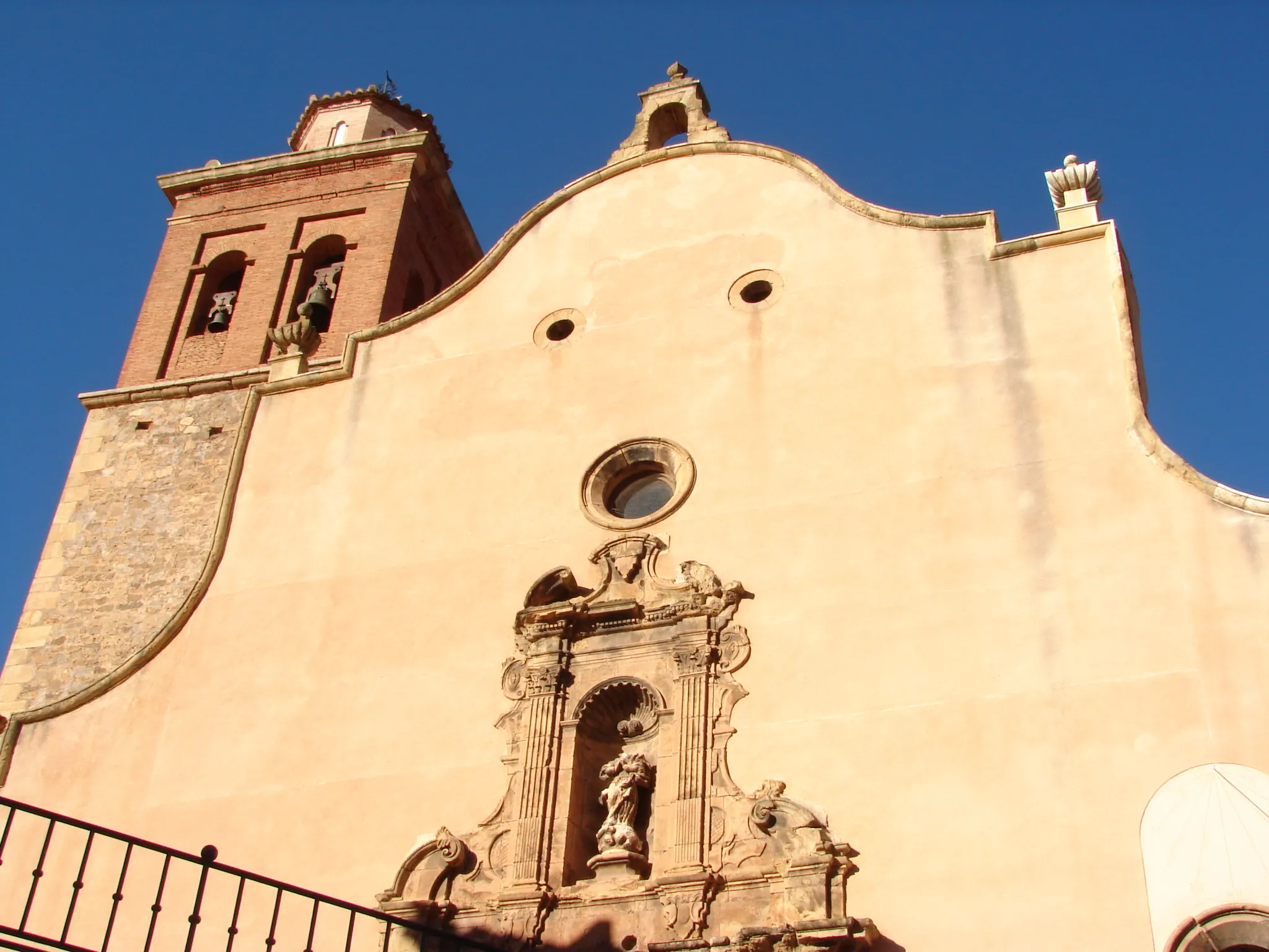 Photo showing: Se trata de una iglesia construida en el s.XVIII siguiendo el estilo
barroco clasicista y concretamente el modelo jesuítico.
Consta de tres naves, de las cuales es mayor la central, mientras
que las laterales son el resultado de la comunicación de
las profundas capillas laterales. Además cuenta con un crucero
no marcado en planta, cabecera recta flanqueada por dos
sacristías simétricas y coro alto a los pies.
La nave central y los brazos del crucero se cubren con bóvedas
de cañón con lunetos separadas por arcos fajones de
medio punto que apoyan en una potente cornisa, mientras que
las naves laterales se cubren con bóvedas de arista y el crucero
con cúpula semiesférica sobre pechinas.
La decoración del interior es típicamente barroca, sin embargo
al exterior destaca la sobriedad de la fábrica de mampostería.
La fachada presenta un remate mixtilíneo y una portada barroca
de dos cuerpos, de los cuales el superior conserva una
hornacina con una imagen de la Inmaculada bastante deteriorada.
En el flanco occidental se alza la torre, que presenta un primer
cuerpo de mampostería, planta cuadrada y gran altura y un
segundo de ladrillo, planta cuadrada y rematado por un tercer

cuerpo octogonal de reducidas dimensiones.