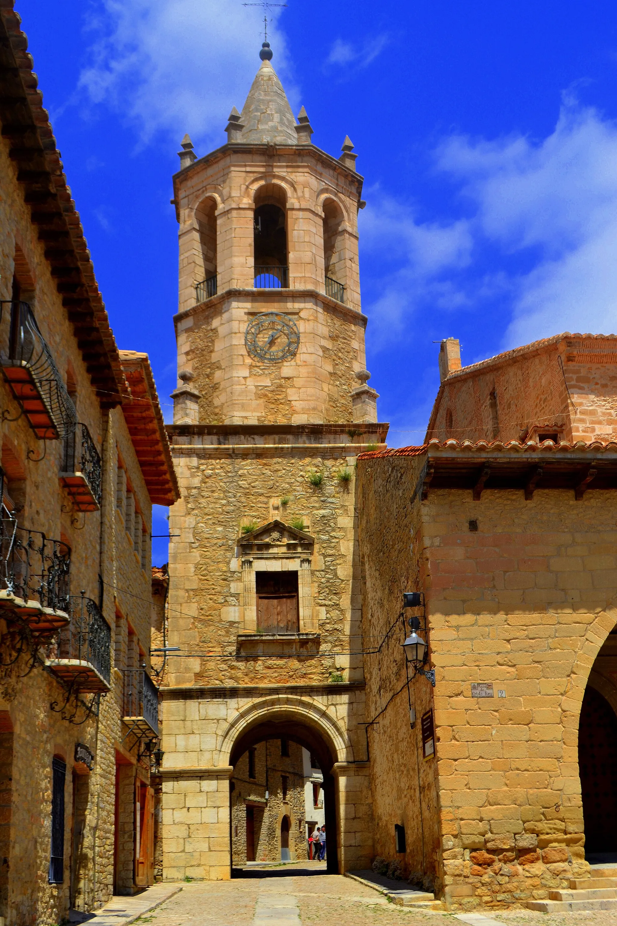 Photo showing: Fue construida según el modelo de la basílica del Pilar, al igual que otros templos de la comarca del Maestrazgo, como la Iglesia de Allepuz y la Iglesia de La Cañada de Benatanduz, y es una de las parroquiales de mayor embergadura de la Provincia de Teruel
En la misma ubicación se encontraba la antigua iglesia parroquial gótica de la cual solo queda el los tres arcos apuntados del pórtico y una pequeña portada a los pies de la iglesia actual.
A partir de 1664, se destruyó la antigua iglesia medieval para construir otra de la cual se conservan únicamente, integradas en el templo actual, la torre y la portada lateral, que hoy es el acceso principal.
El templo barroco fue construido entre 1730 y 1745, según un proyecto de Antonio Nadal.
Sufrió las consecuencias de la Guerra Civil, durante la cual fueron destruidos la mayor parte de sus suntuosos retablos, quedando únicamente testimonio fotográfico, si bien se salvaron las pinturas murales y algunos fragmentos de retablos.
Alguna de estas capillas pertenecían a las familias nobles de la comarca como los Osset cuya heráldica se conserva en la embocadura de una de ellas.
Es una obra barroca de grandes dimensiones, con una distribución en tres naves de similar altura, según el modelo clasicista extendido en Aragón a partir de la construcción de la basílica del Pilar. Está realizada en mampostería.
Por el exterior destaca, en el lado de la Epístola, un pórtico abierto a la plaza Mayor formado por tres grandes arcos apuntados de sillería, pertenecientes a la anterior iglesia gótica. La portada es sencilla, con arco de medio punto enmarcado por un orden de pilastras bajo frontón.
En el interior, las naves, de idéntica altura, están separadas por gruesos pilares acanalados que sustentan arcos de medio punto. La nave central está cubierta mediante bóveda de cañón con lunetos, mientras que las laterales lo están con bóvedas de arista. El crucero presenta una cúpula elíptica sobre tambor y pechinas decoradas con relieves de los evangelistas y rocalla; al exterior aparece como un volumen octogonal de ladrillo.
Dispone asimismo de una amplia girola tras la cabecera y de coro a los pies, en el penúltimo tramo de la nave central, decorado con pinturas que representan a San Braulio, San Francisco Javier y otros personajes sagrados.

La torre, situada a los pies, da paso a una calle, adoptando la misma solución que las iglesias de la capital turolense. De planta cuadrada, presenta en su parte superior dos cuerpos octogonales y un remate piramidal.