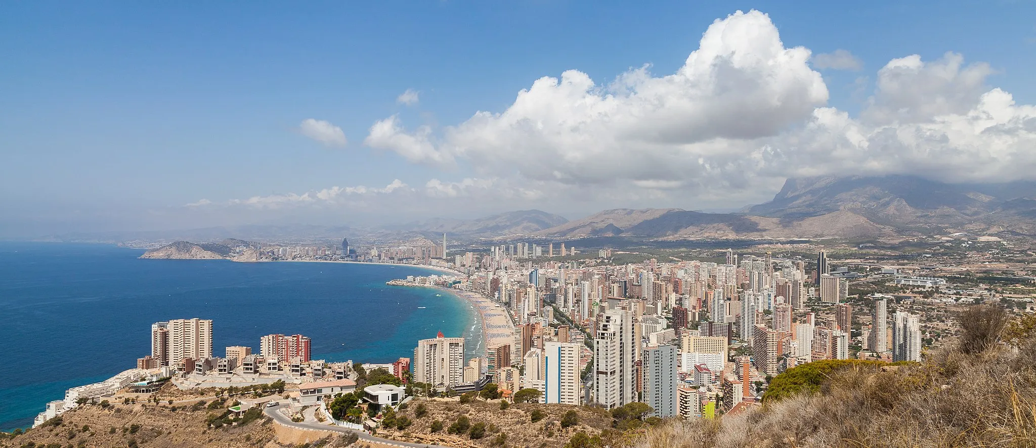 Photo showing: View of Benidorm, turistic capital of the Costa Blanca (literally White Coast) in Land of Valencia, Spain. The shot was taken from the Cross of Benidorm, located on the summit of the Sierra Helada. Benidorm, is a town with 73,000 inhabitants throughout the year but with a peak of over half a million in the summer season. It's the third town with the most concentration of tall buildings in Europe, after London and Milan, whereas in Spain, Benidorm is positioned third, behind Barcelona and Madrid in the total number of skyscrapers. Nevertheless, Benidorm has the most high-rise buildings per capita in the world.
