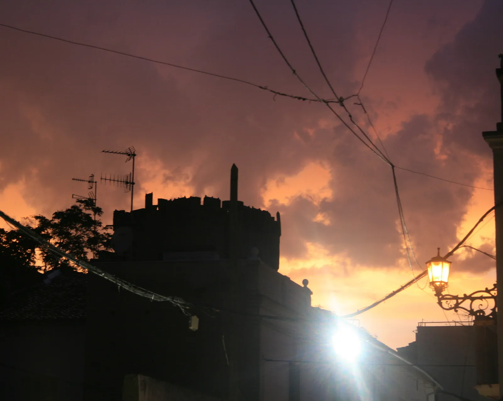 Photo showing: Coria sunset after rain.  Sun is setting behind black silhouette of castle; and it lights heavy dark clouds from below