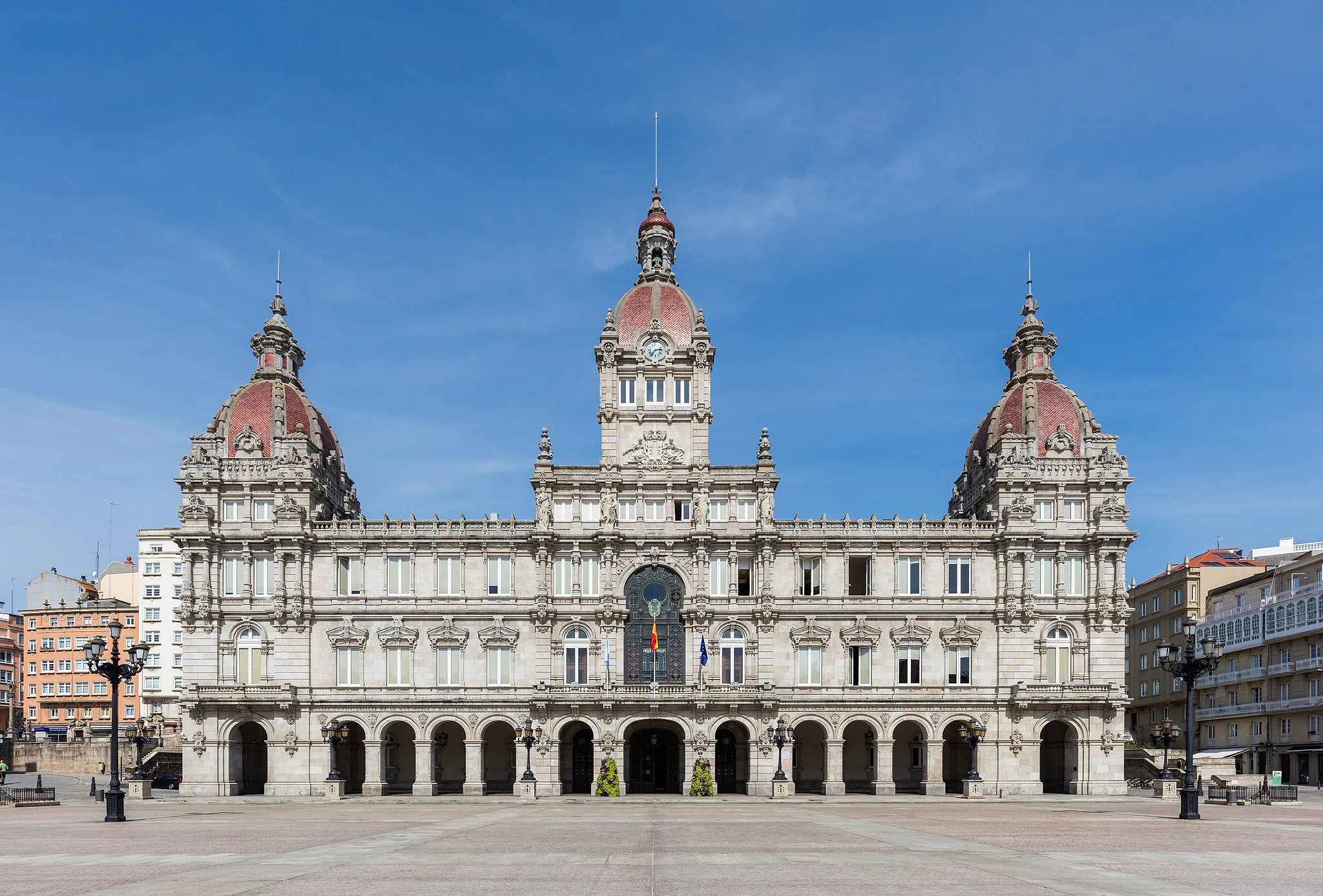 Photo showing: Front view of the City Hall, also called the Municipal Palace, of A Coruña in Galicia, Spain, during the blue hour. The modernist building is located in the María Pita Square in the center of the city and was built between 1908 and 1912 following a design of Pedro Ramiro Mariño. It was inaugurated in 1927 by king Alfonso XIII. The facade is 64 metres (210 ft) wide and has 43 windows. The ground area of the building is 2,300 m2 (24,757.0 sq ft). The four statues on the third floor represent the four provinces of Galicia — A Coruña, Lugo, Orense and Pontevedra. Over them, in the middle, is the coat of arms of the city. The two dames flanking it symbolize Peace & Industry and Work & Wisdom. The tower in the middle accommodates the clock and the bells, which are made of bronze and tin and weigh 1,600 kilograms (3,527 lb).
