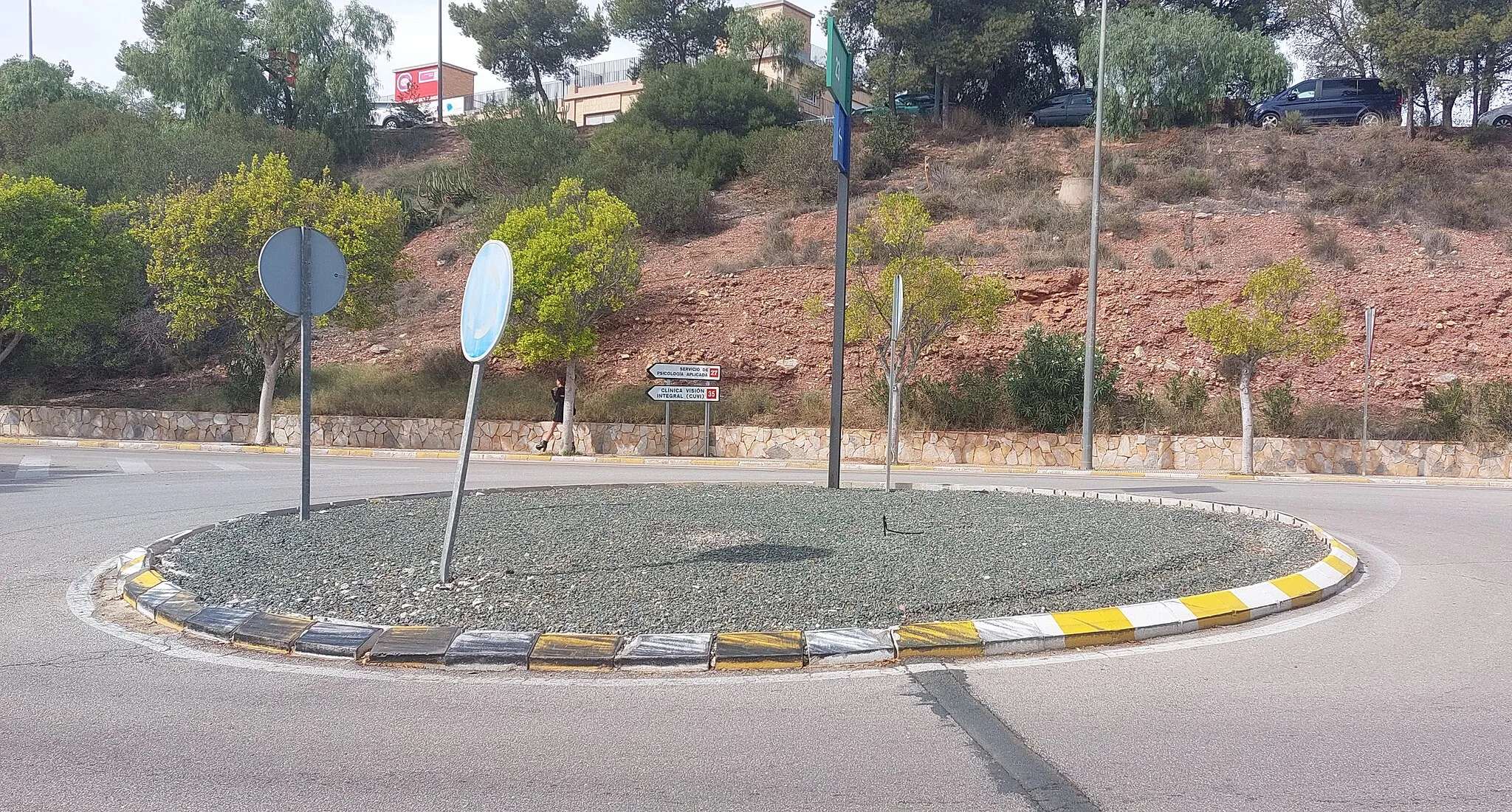 Photo showing: Photograph of the current state of the roundabout at the Faculty of Medicine, University of Murcia; featuring a bare roundabout covered with green gravel and having three exits. Previously, it housed a car split in half adorned with plants.